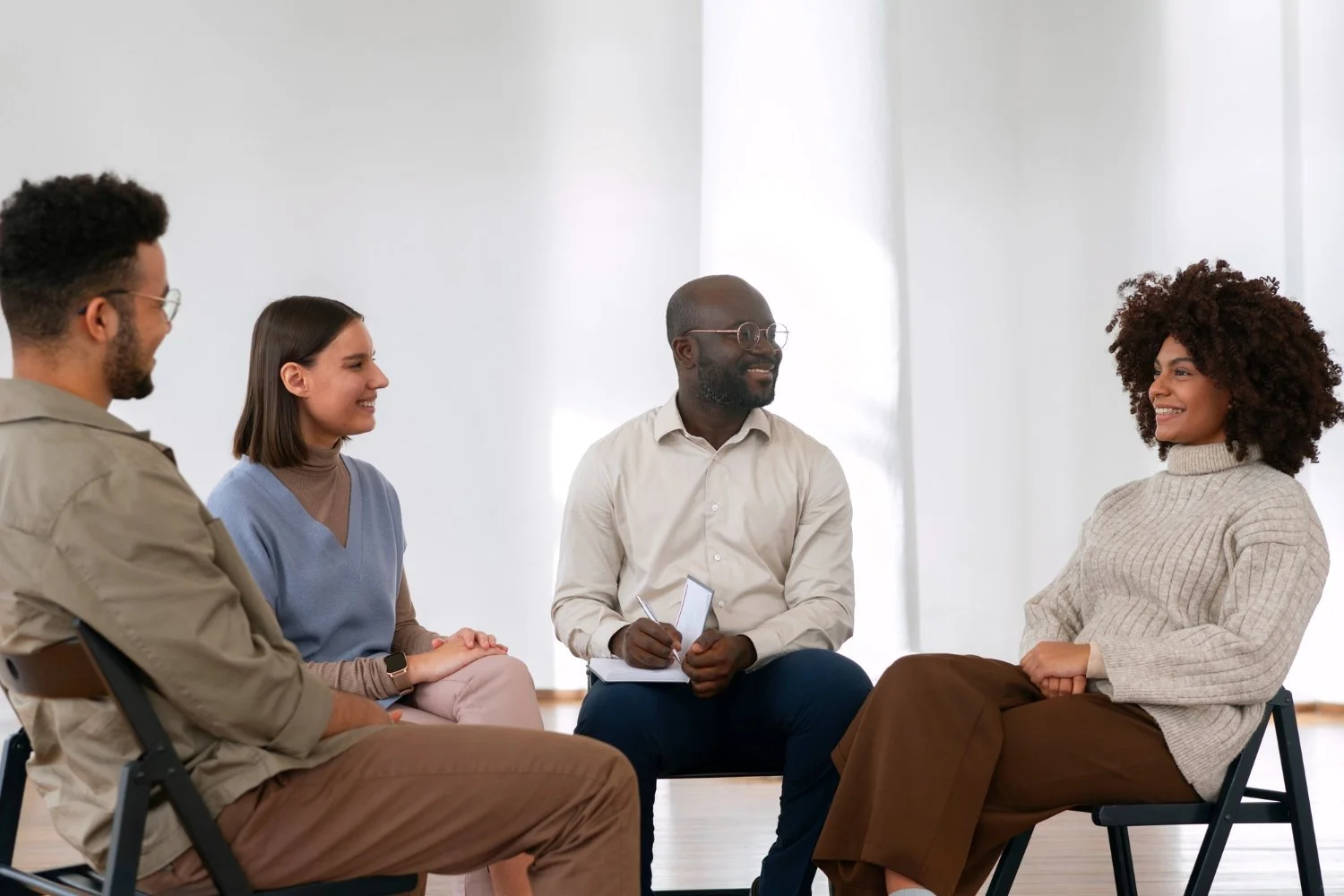 People sitting in a circle during a group therapy session or support group meeting in a bright room.