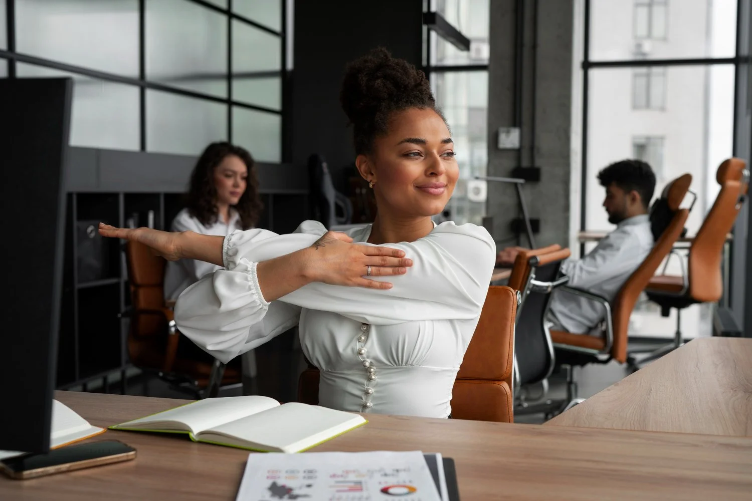 A woman stretching her arm across her chest in an office setting with two other coworkers working at desks in the background.