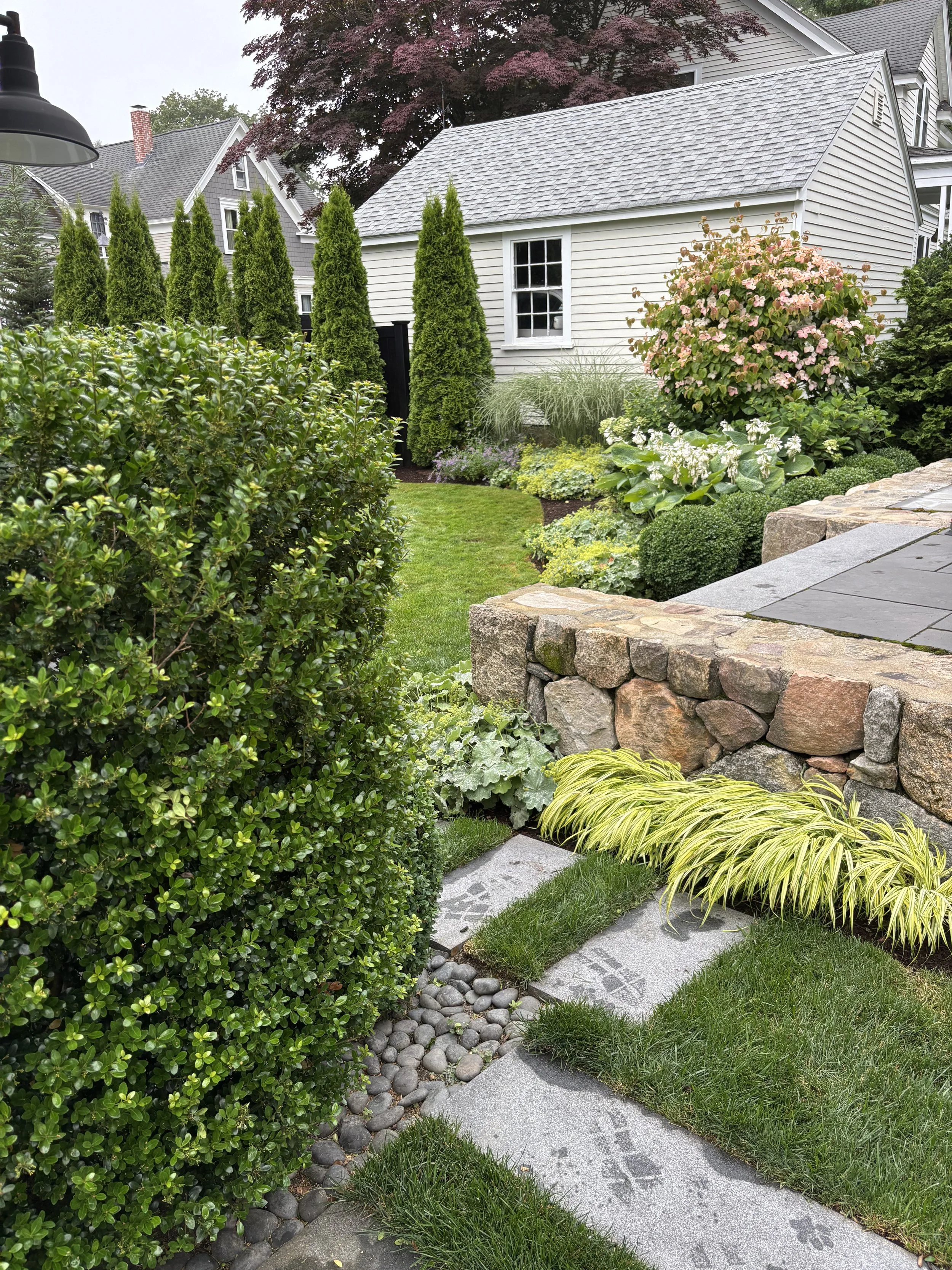 A raised patio created by fieldstone wall and pavers is flanked by gardens and lawn area.