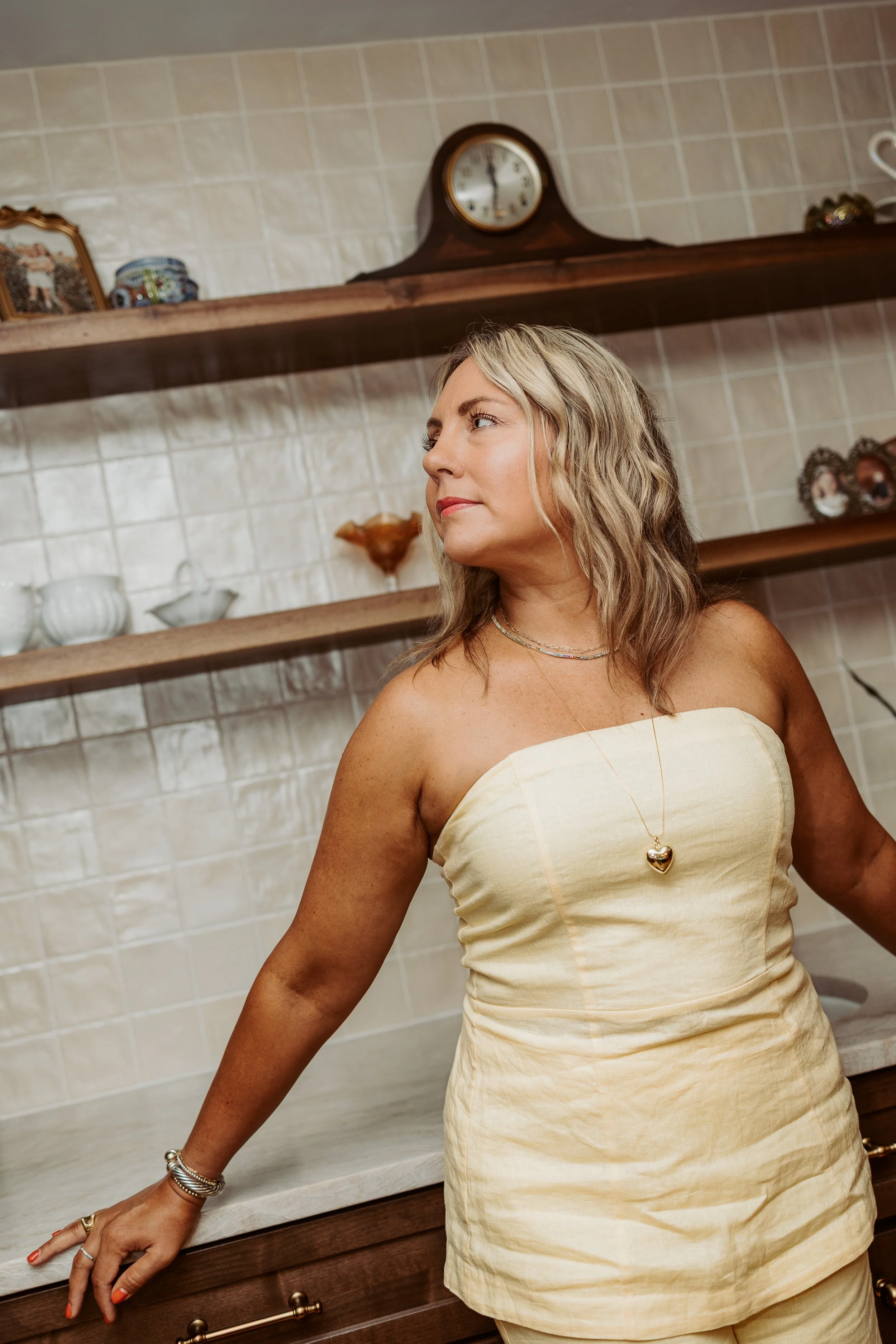 A woman with wavy blonde hair wearing a strapless, cream-colored dress with a heart-shaped necklace, standing in a kitchen with wooden shelves and decorative items, including a clock, plates, and framed photographs.