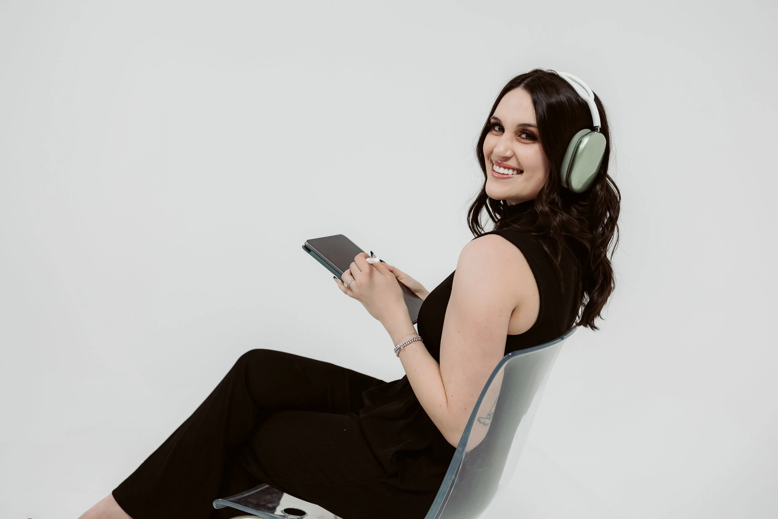 A woman with dark brown hair smiling, sitting on a clear plastic chair against a plain white background, wearing a black sleeveless top, black pants, a bracelet, holding a smartphone, and wearing large over-ear headphones.