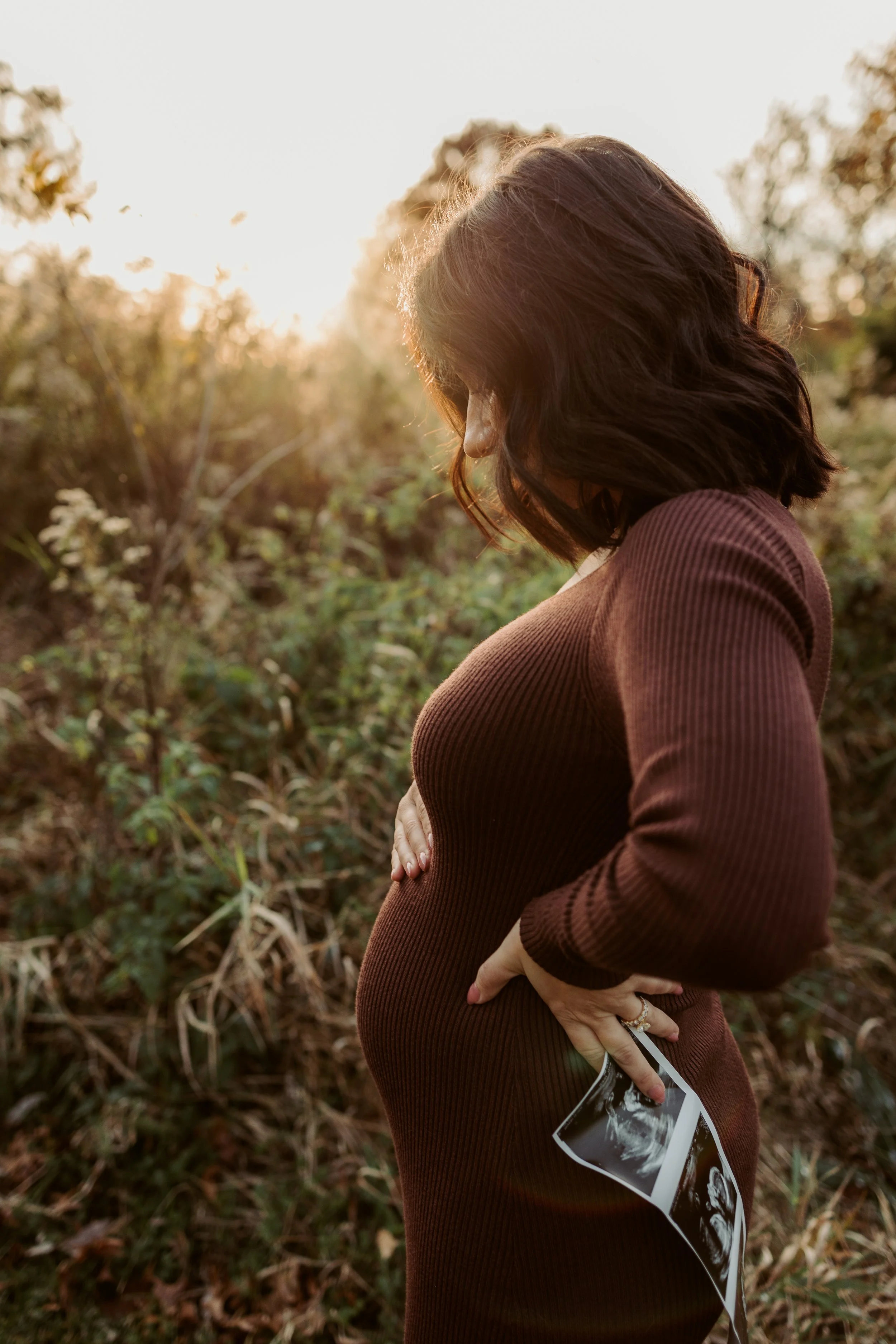 Pregnant woman in a brown dress holding ultrasound pictures, standing outdoors during sunset or sunrise.