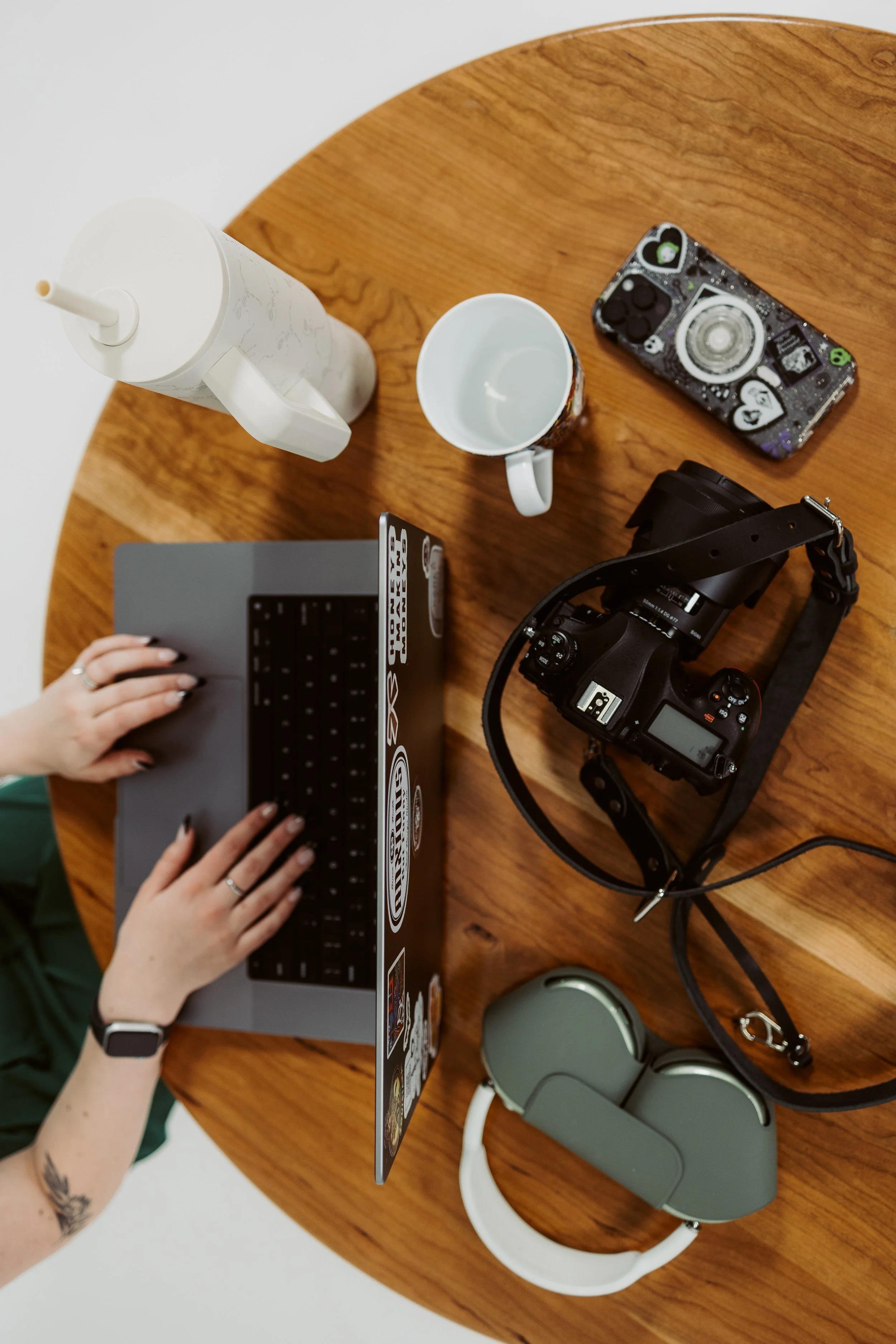 A wooden table with a white water bottle, a white mug, a black and white camera, a gray headset, a portable speaker, a laptop, and a person typing on a keyboard.