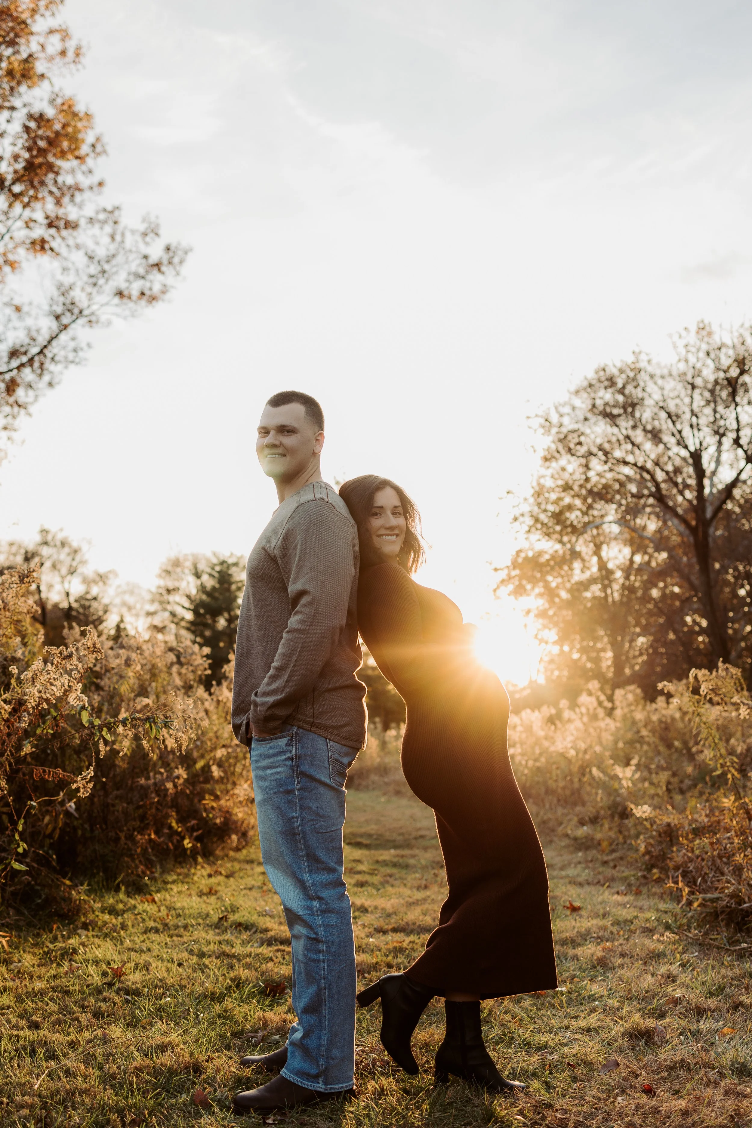 A couple stands back to back in a field with autumn trees in the background, smiling at the camera during sunset.