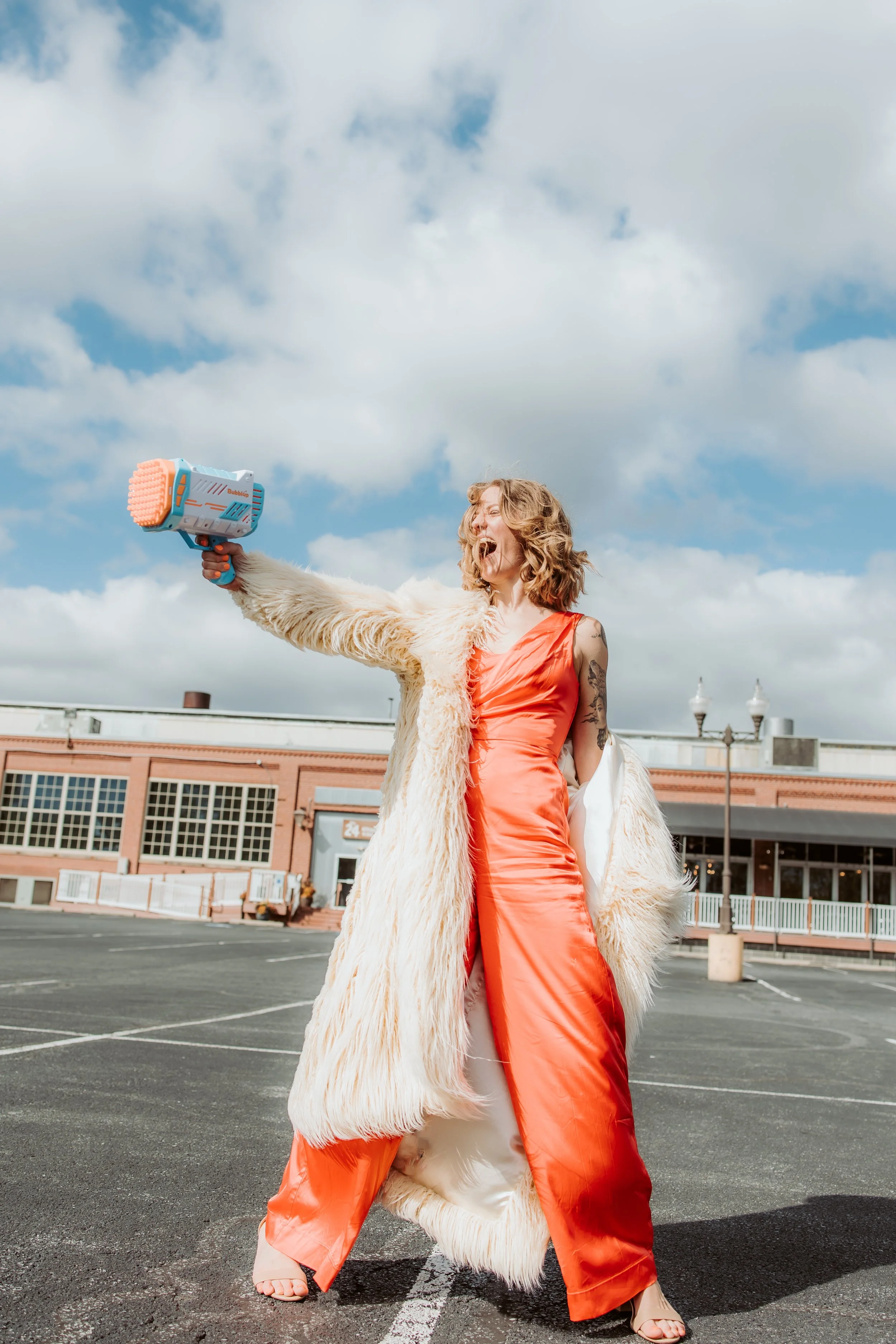 Woman in an orange satin jumpsuit and beige heels holding a toy blaster, standing outdoors in an empty parking lot, amusingly pretending to shoot, with a cloudy sky overhead.