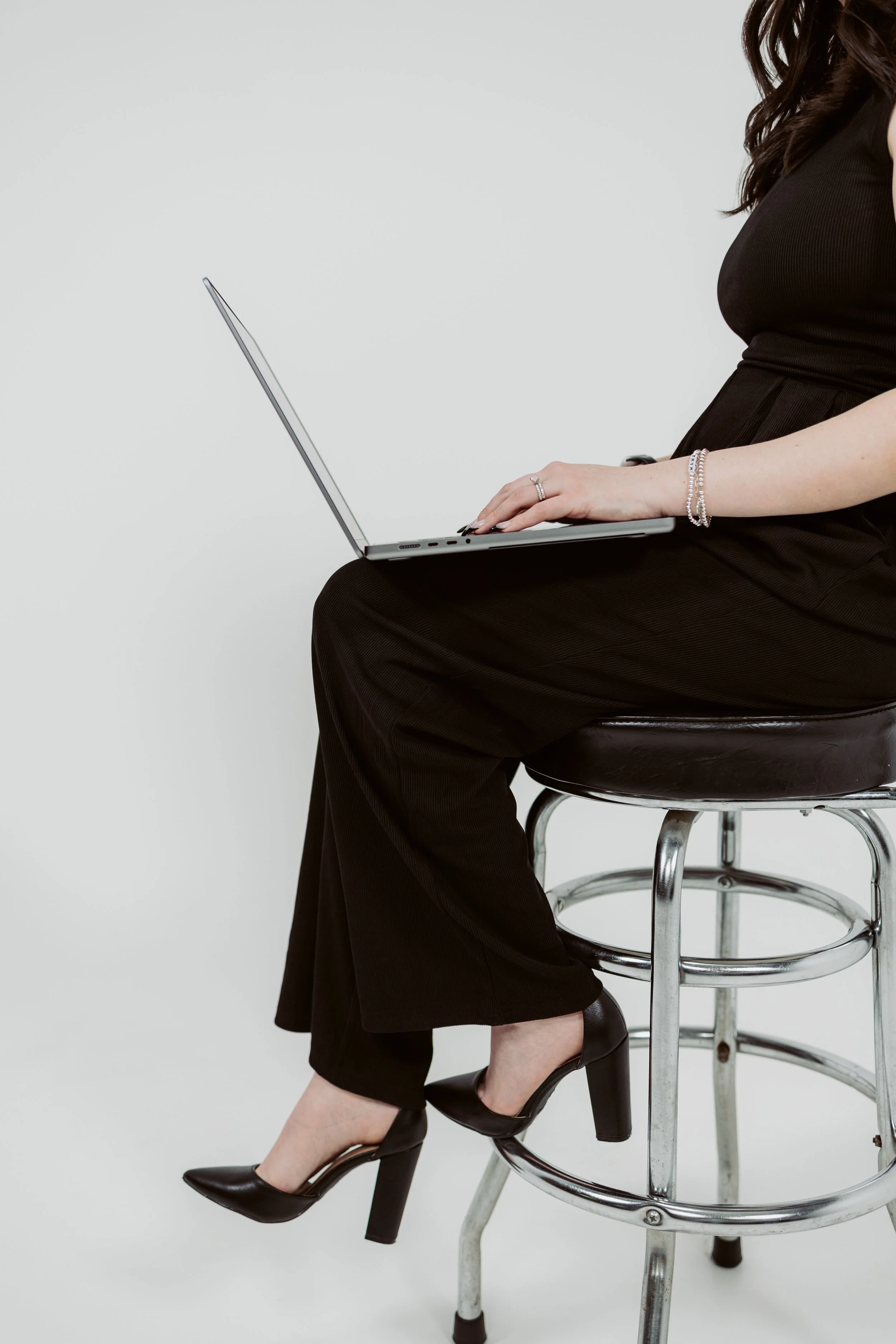 A woman in a black dress and heels sitting on a metal stool, holding a silver laptop on her lap.