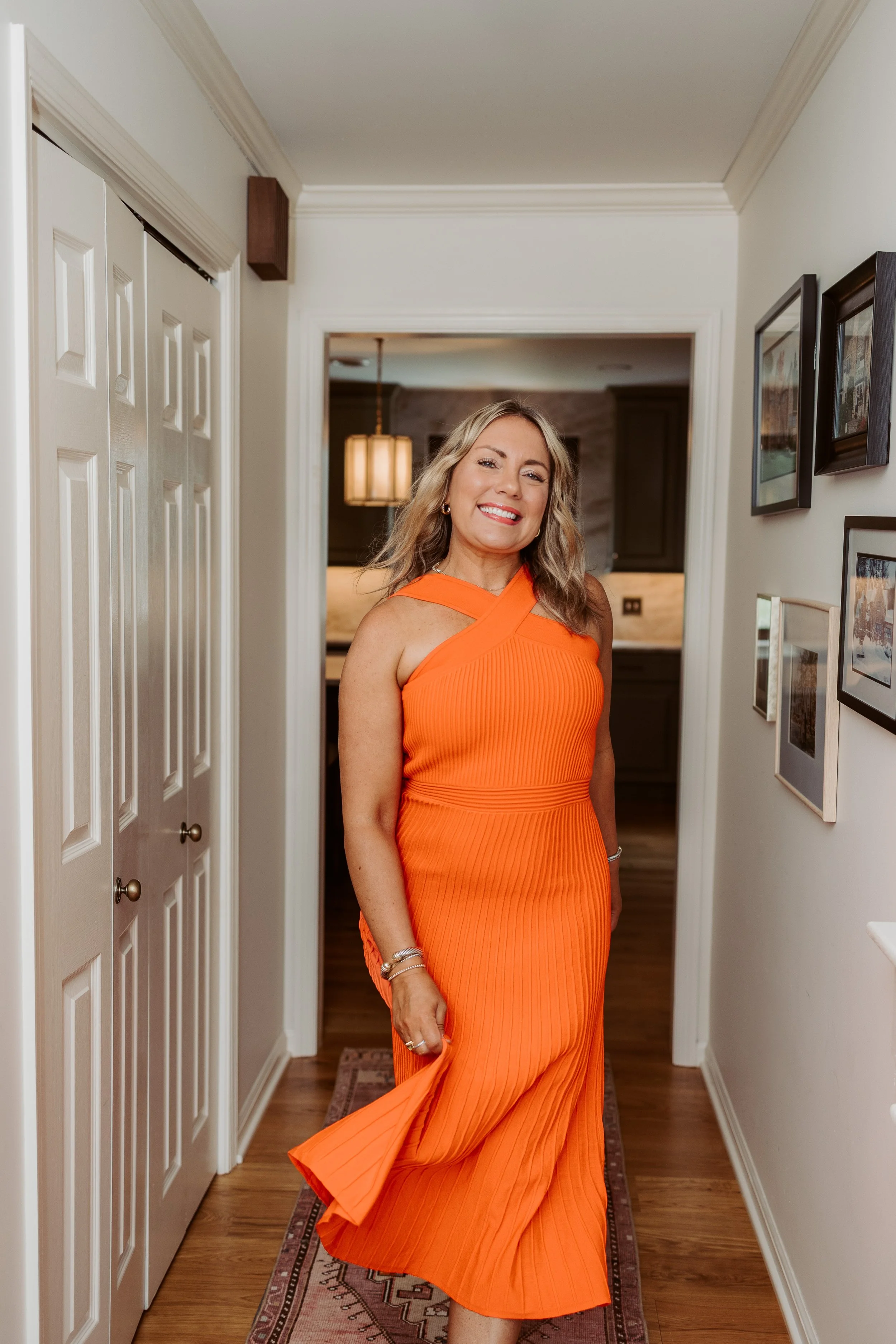 A woman with wavy blonde hair is standing in a hallway wearing an orange dress with a pleated skirt and a geometric neckline, smiling at the camera.