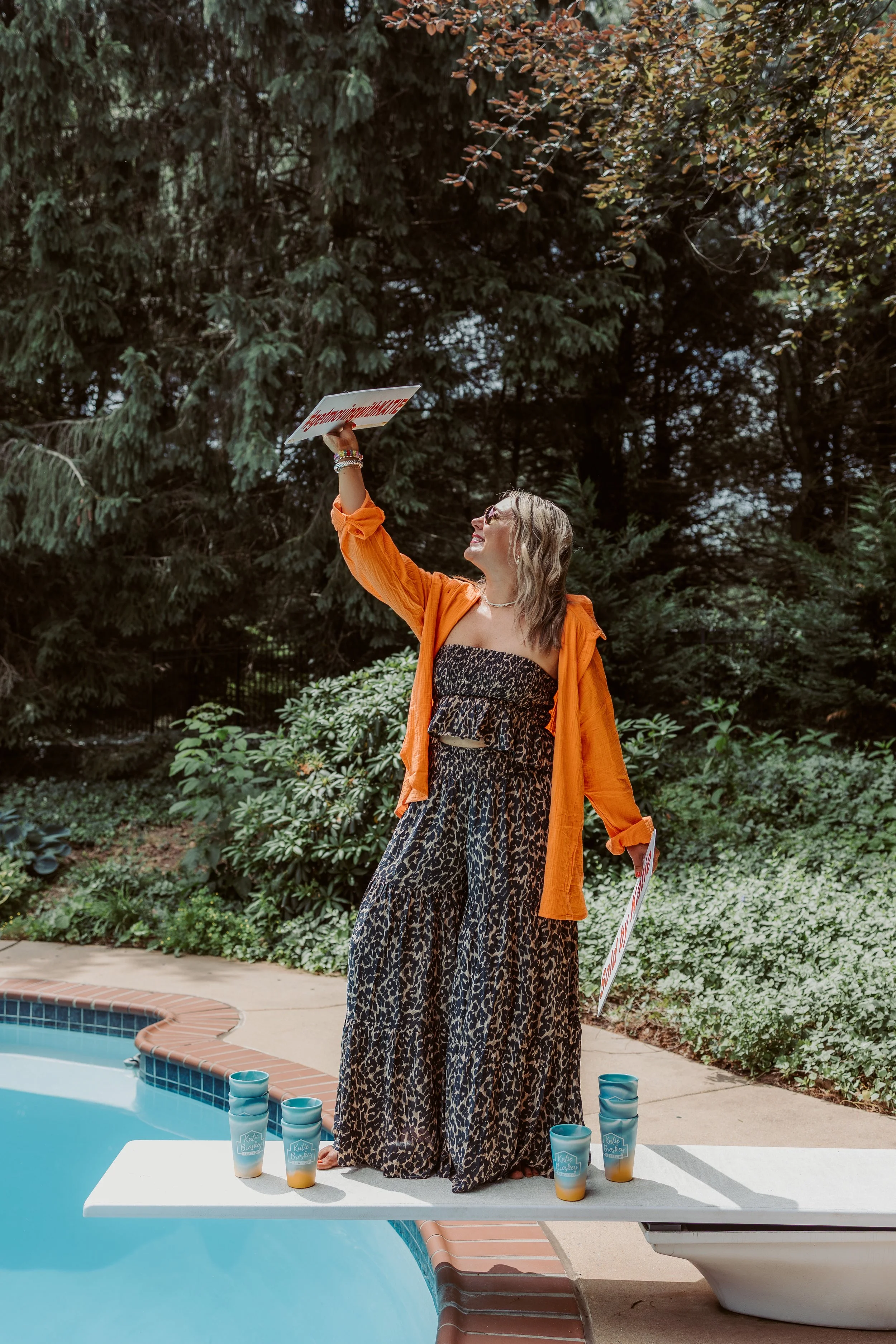 A woman in a leopard print maxi dress and orange cover-up stands on a diving board by a swimming pool, holding a sign and smiling. There are cupcakes arranged on the board, and greenery and trees in the background.