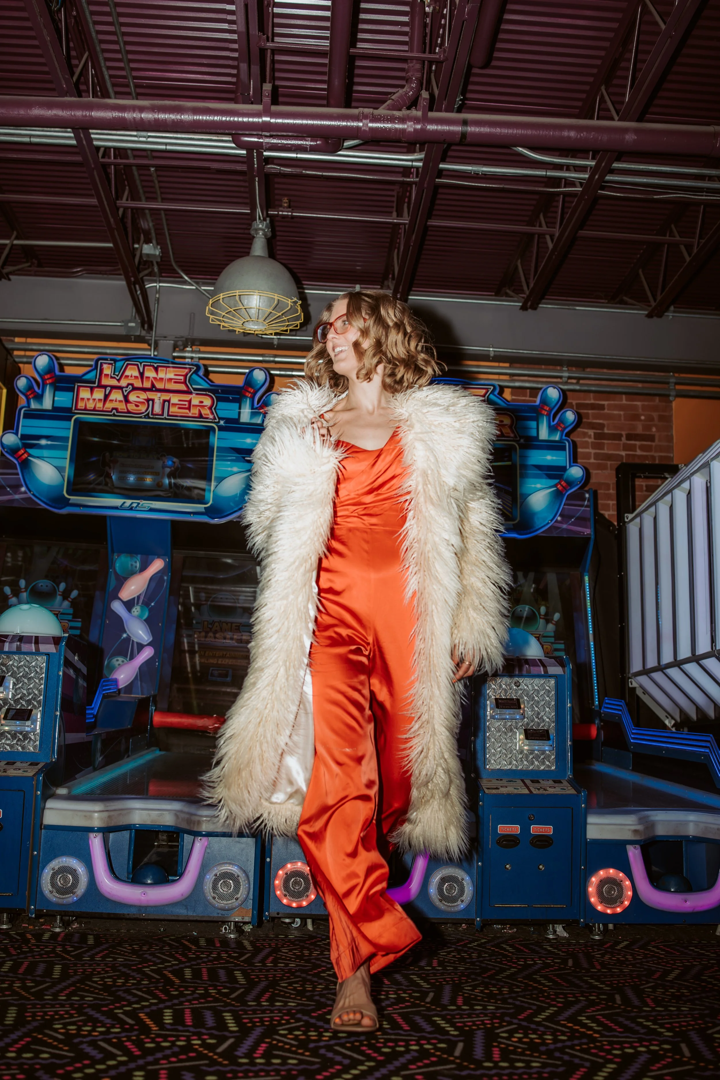 Woman with curly hair and glasses standing in front of an arcade game, wearing a luxurious fluffy coat over an orange satin dress.