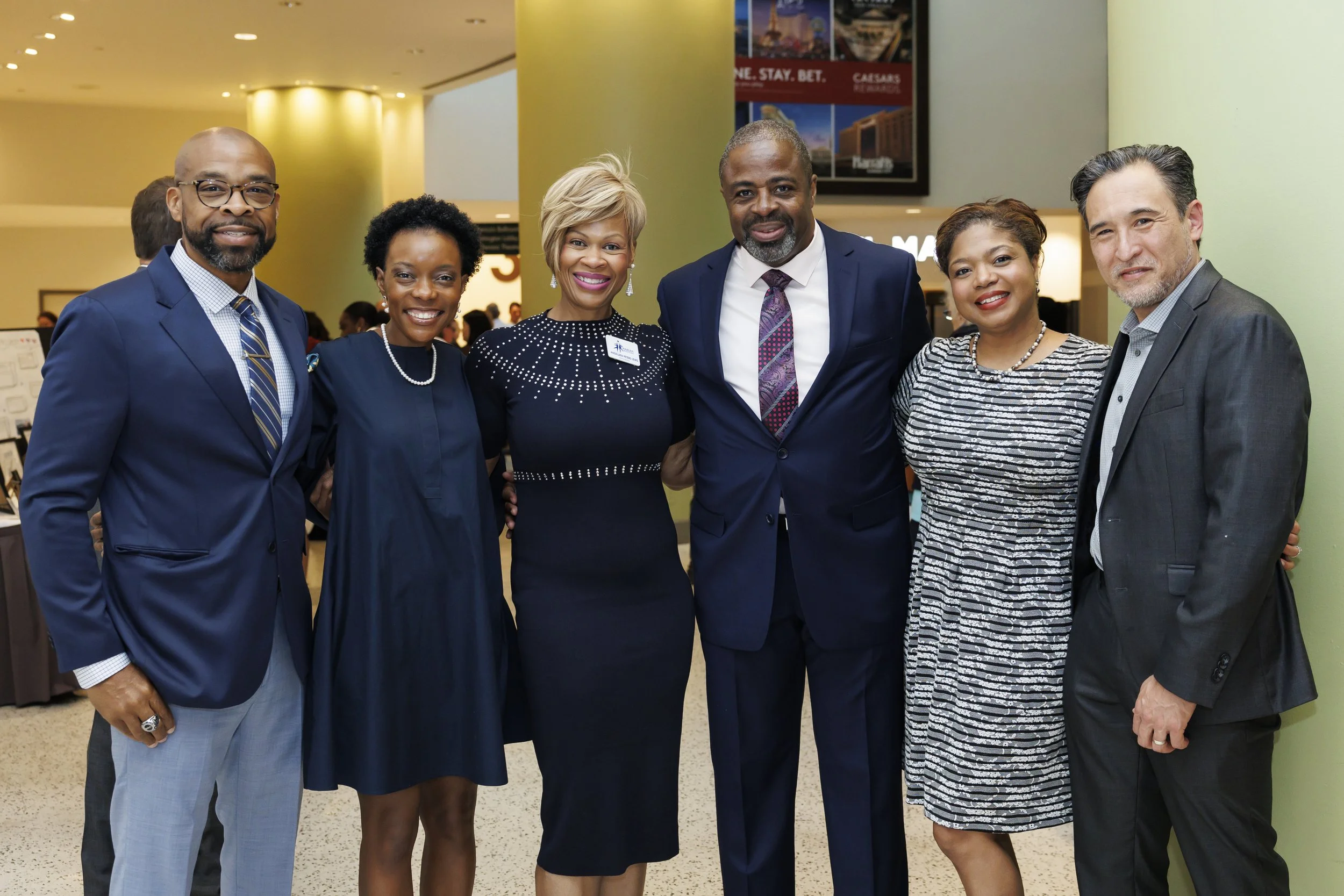A group of six professionally dressed people smiling and posing together in an indoor setting, to support the Marian Magic 2025 fundraiser.