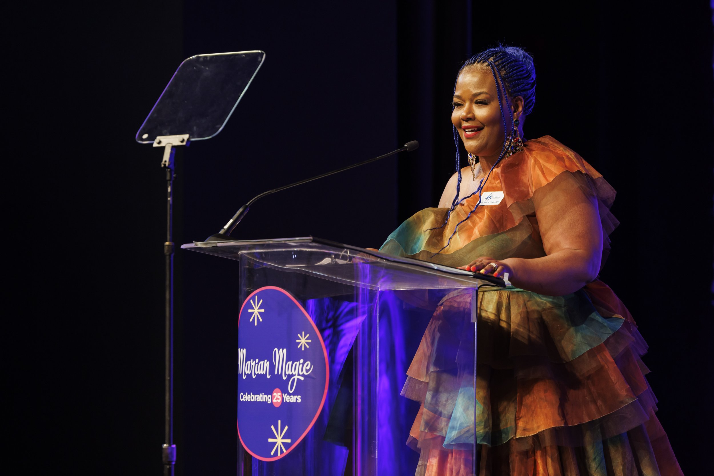A Marian Middle School educator with braided blue hair standing at a podium, speaking into a microphone, during an event celebrating 25 years of Marian Magic, wearing a colorful, multi-layered dress.