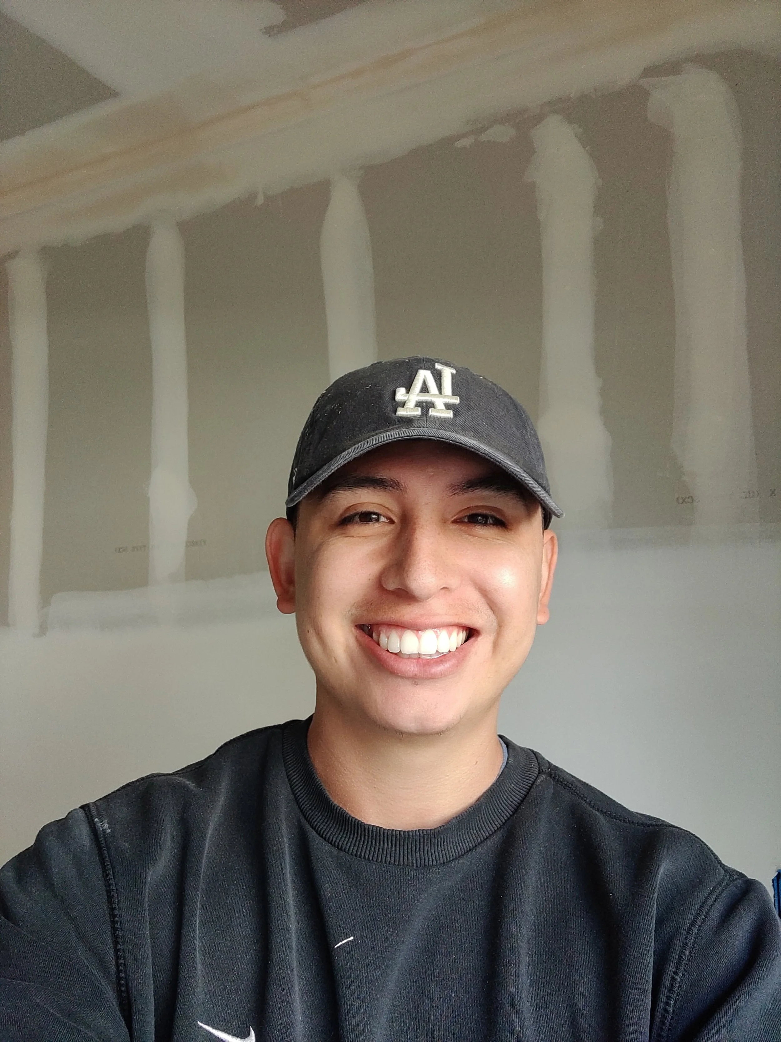 A smiling young man wearing a black Nike sweatshirt and a black Los Angeles Dodgers baseball cap, taking a selfie in front of an unfinished drywall wall with visible joint tape and compound.