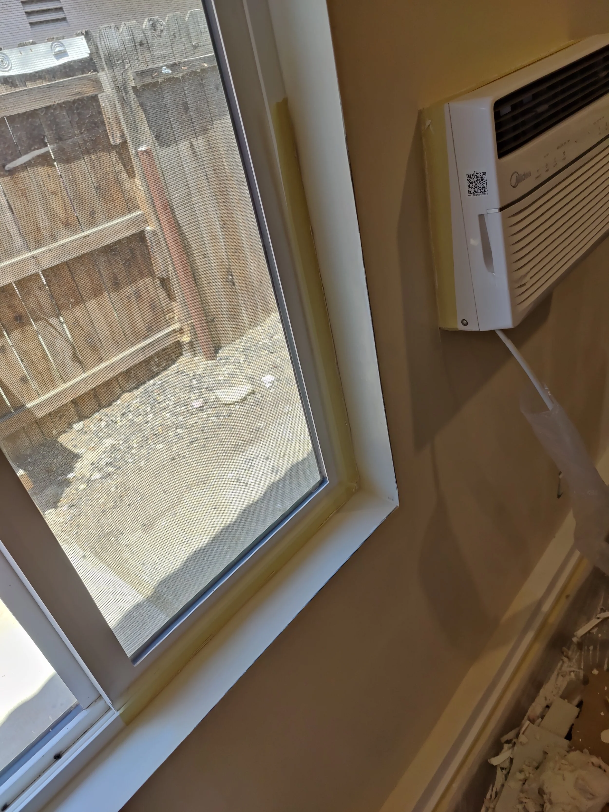 An indoor window with a view of a wooden fence and dirt yard outside, and a white air conditioning unit installed beneath the window.