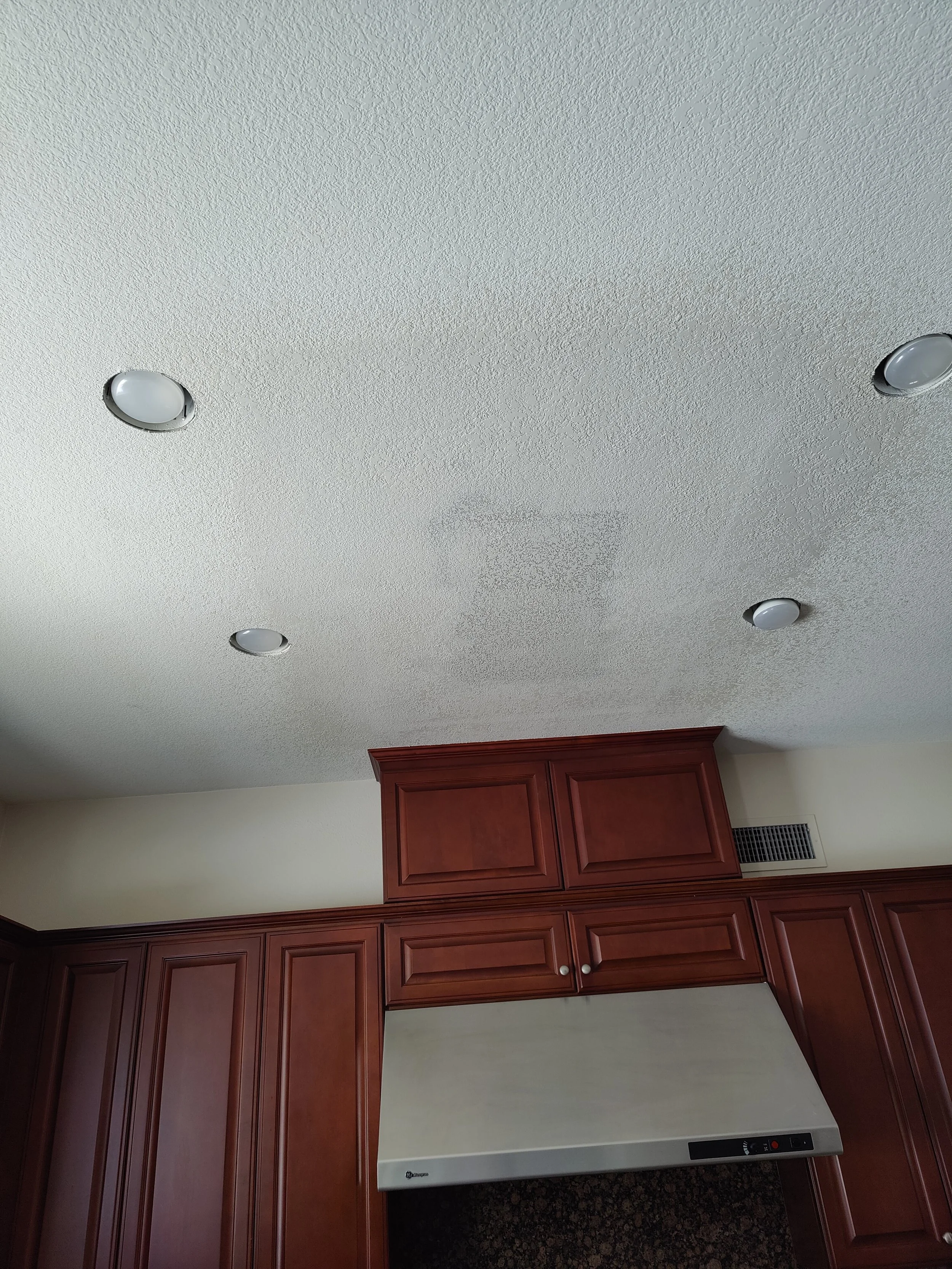 Ceiling with four recessed lights and kitchen cabinets with a vent in the wall.
