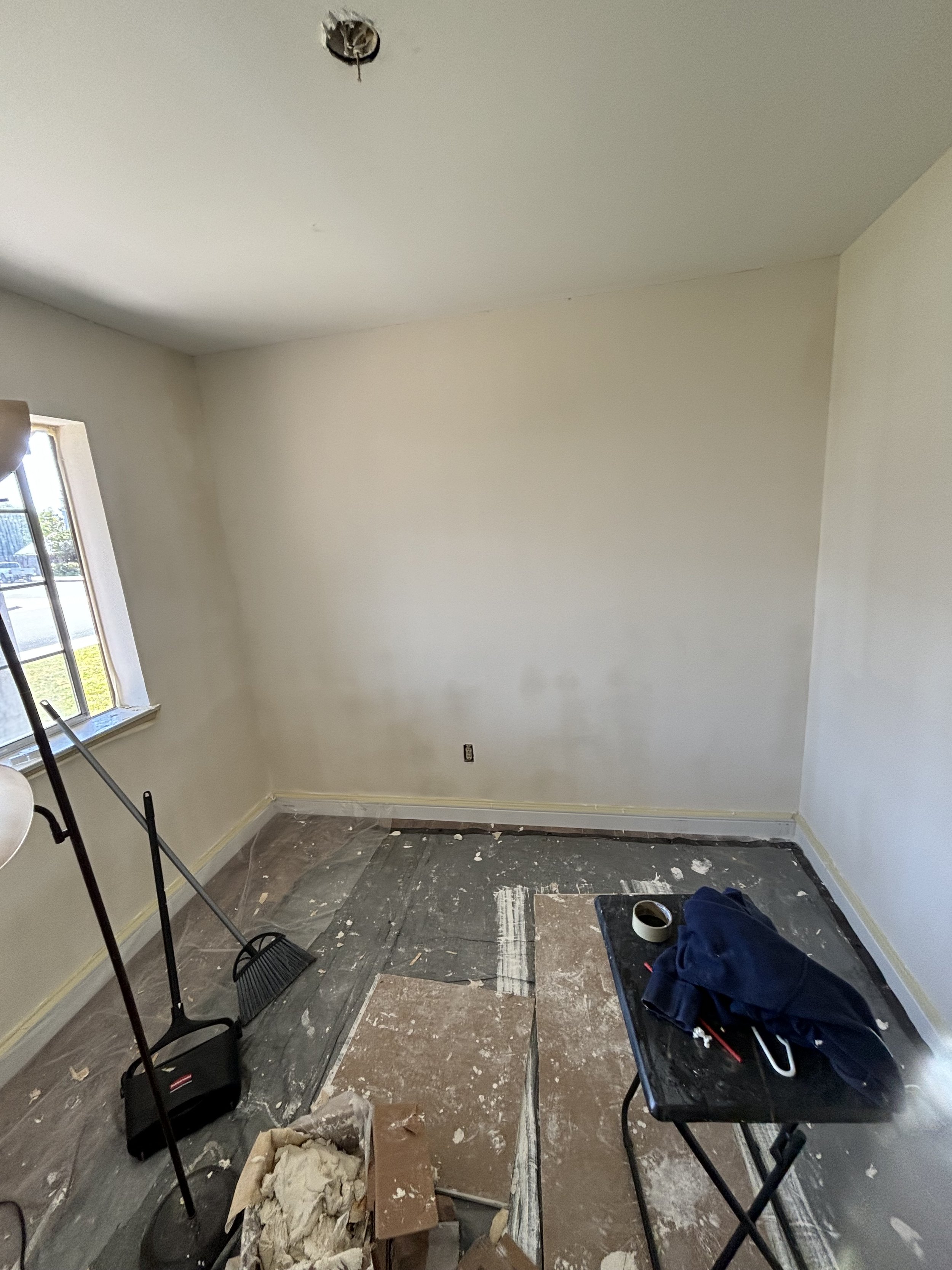 Room under renovation with a window, trash, painting supplies, brooms, tape, and ladders.