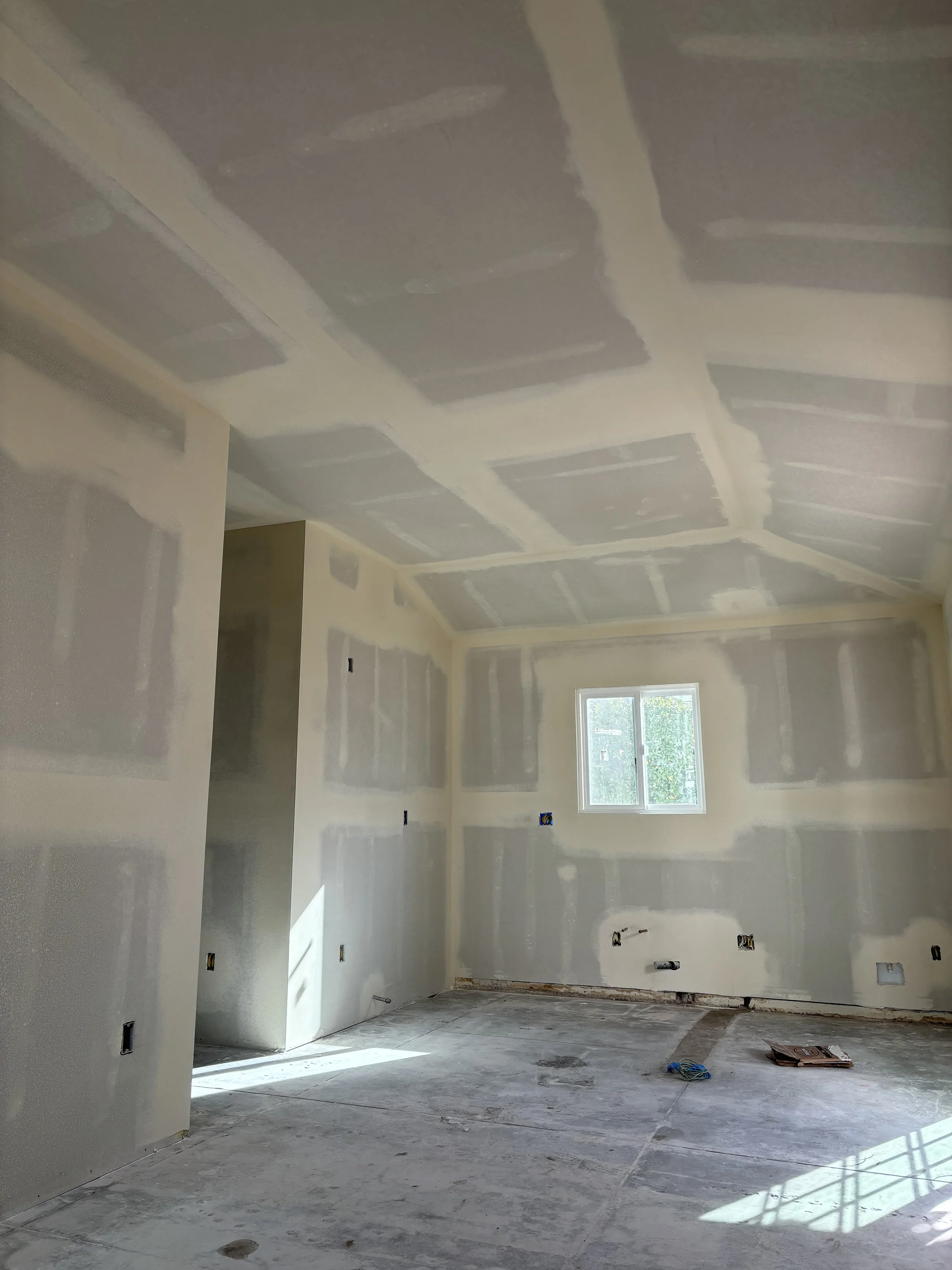 An unfinished room under construction with drywall installed, all seams taped and mudded, and a window letting in natural light.