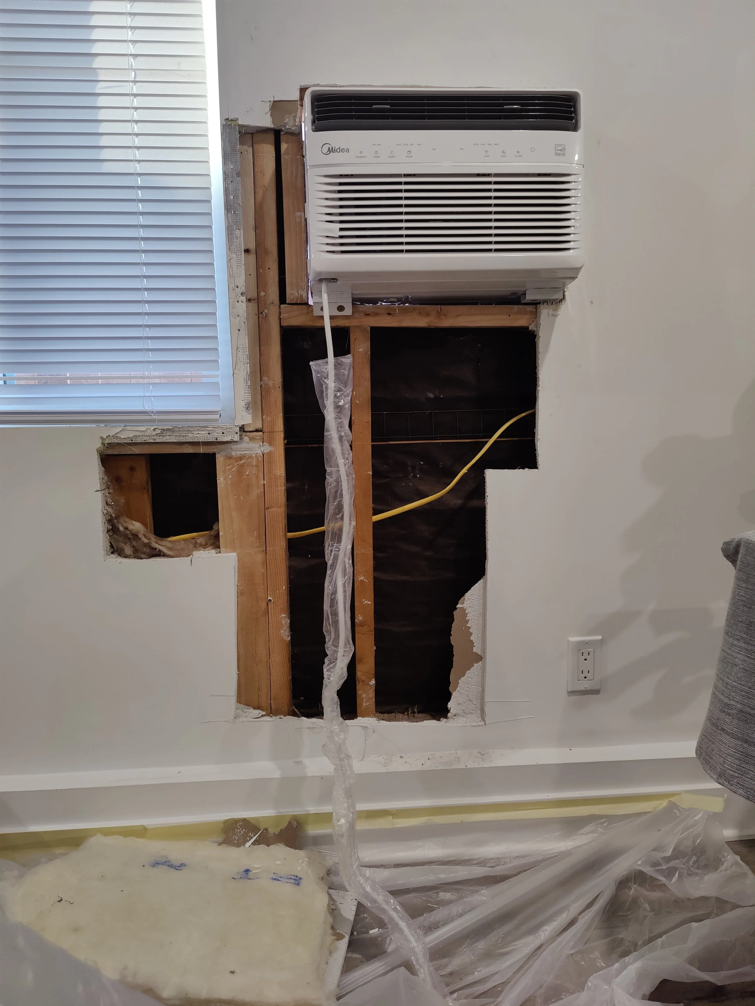Wall with insulation, wooden studs, and an opening where an air conditioning unit is being installed, with plastic sheeting and construction materials nearby.