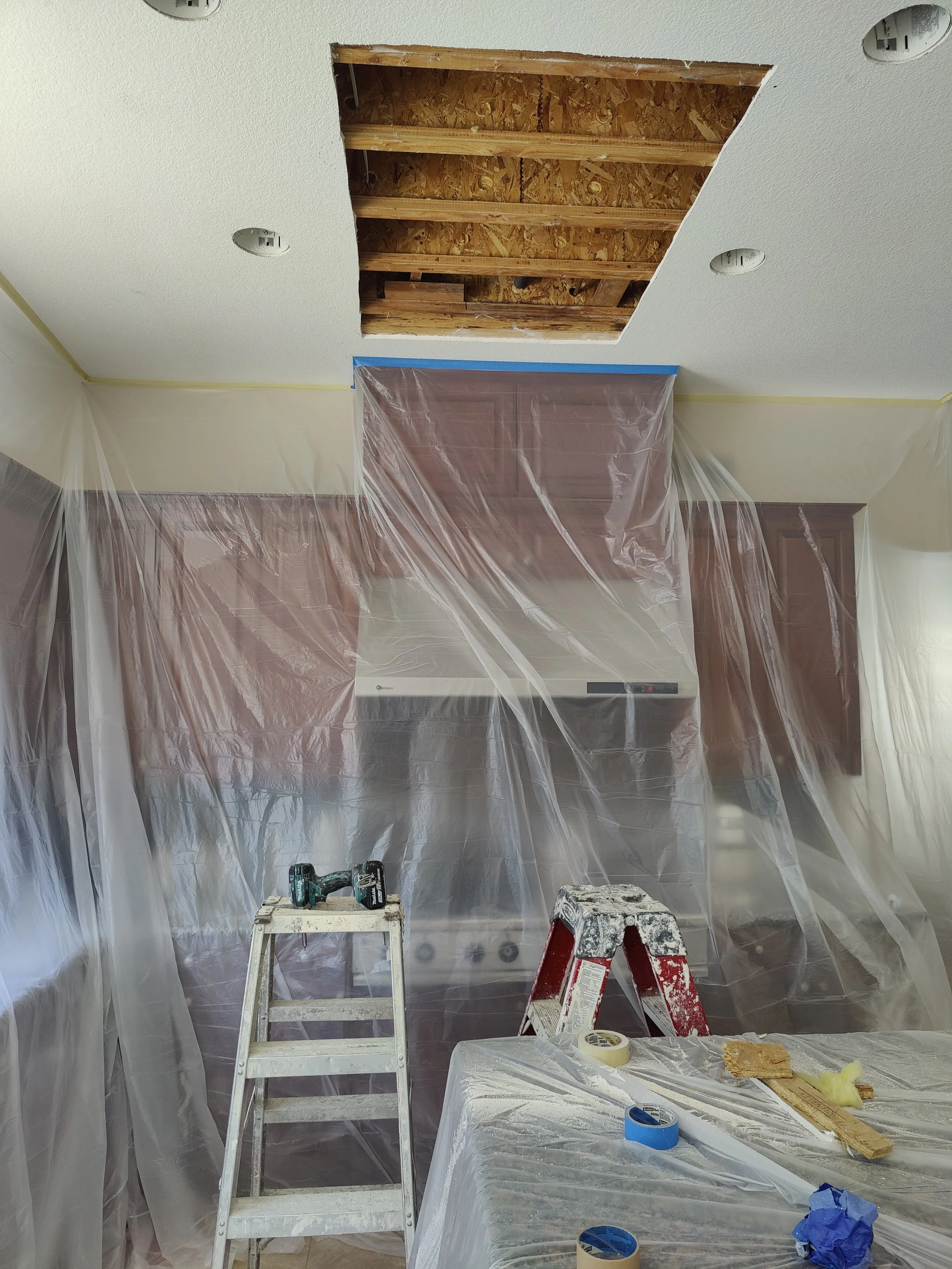 Kitchen ceiling undergoing renovation with a hole exposing wooden beams, surrounded by plastic sheeting, a stepladder, tools, and construction materials.