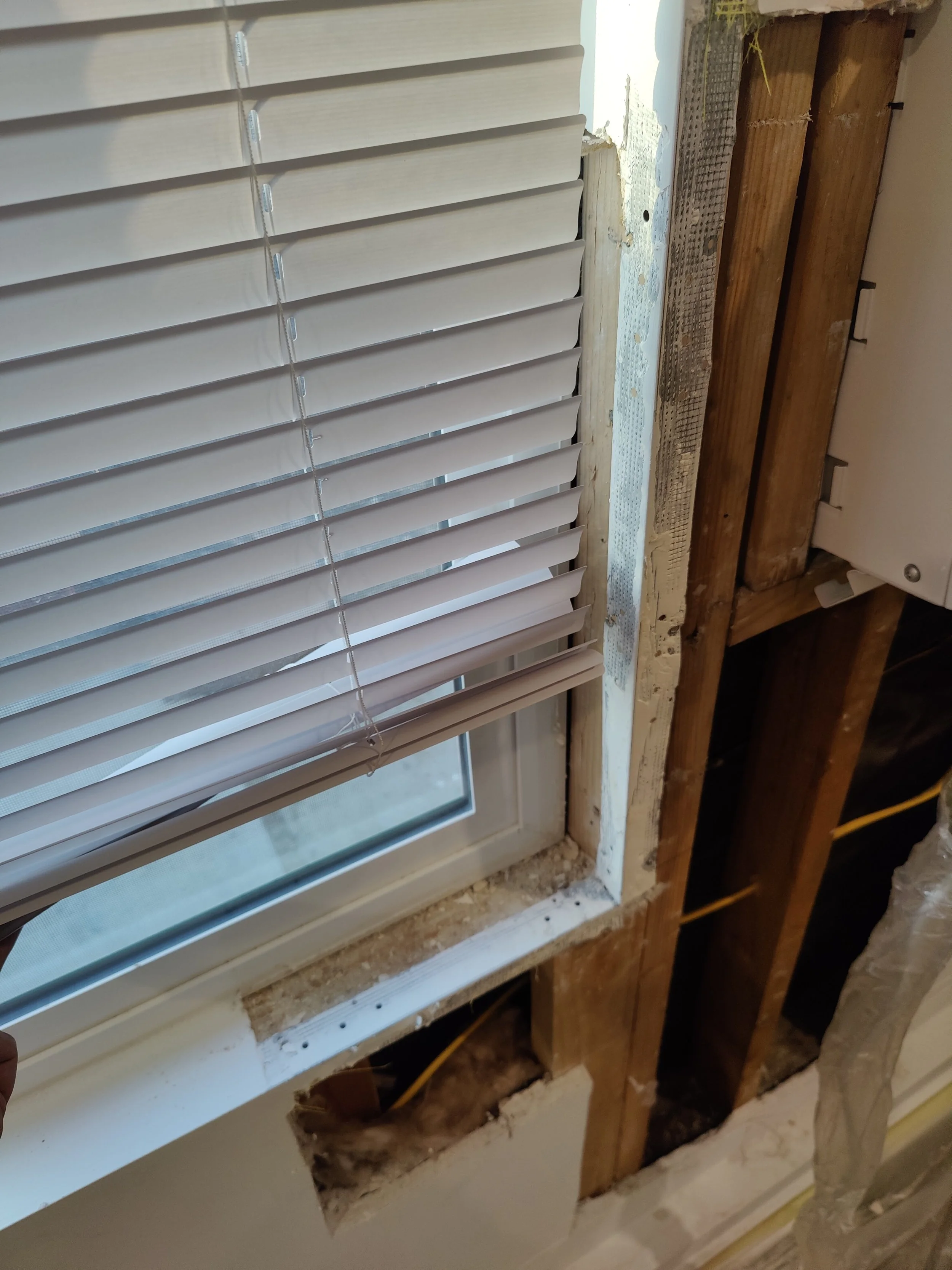 A window with white blinds installed in a wall with exposed wooden framing and unfinished drywall around it under construction.