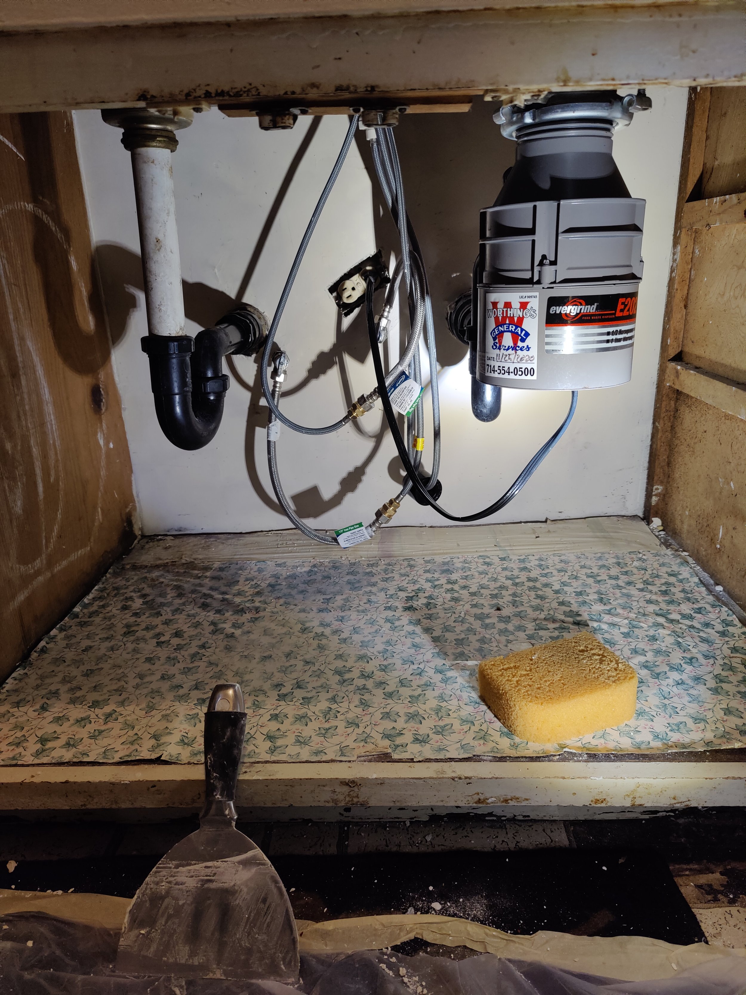 Underneath a kitchen sink showing plumbing pipes, flexible water supply hoses, and a garbage disposal unit with an energy branding label. A sponge lies on a decorative mat in the space, which is partially surrounded by wood panels and wall surfaces.