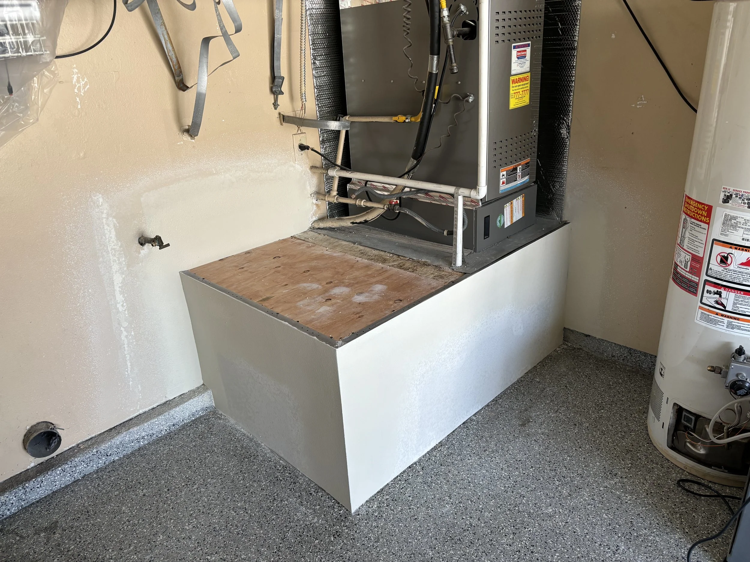 Unfinished laundry room with a water heater on the right, a raised platform with an HVAC unit on top, drywall installed around water heater platform and empty wall plumbing fixtures.