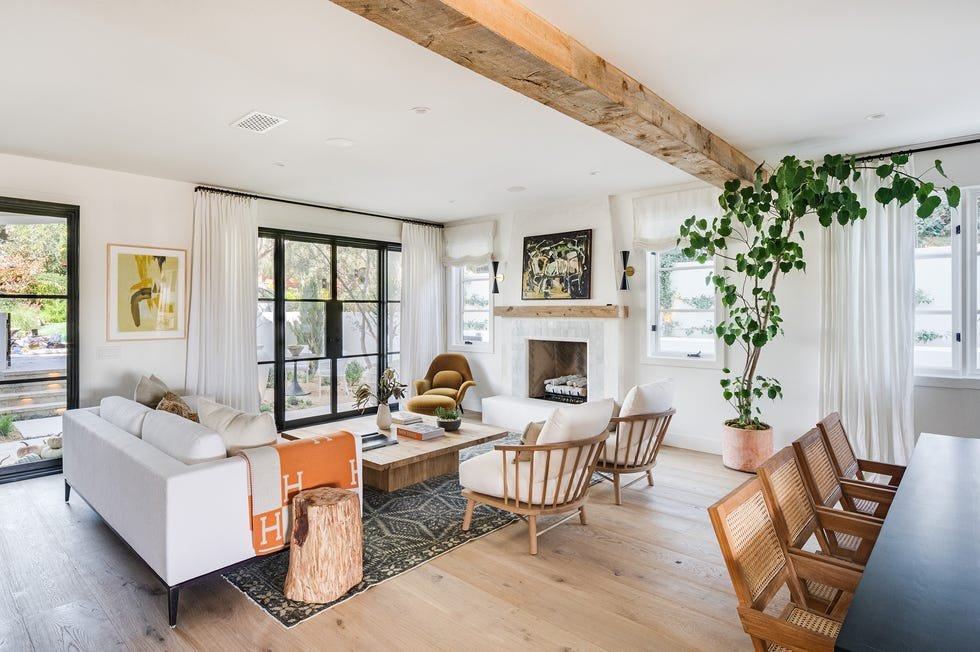 Modern living room with white walls, wooden beams, large windows with white curtains, a white sofa with throw pillows, wooden chairs, a fireplace, artwork on the walls, a large potted plant, and a wooden coffee table.