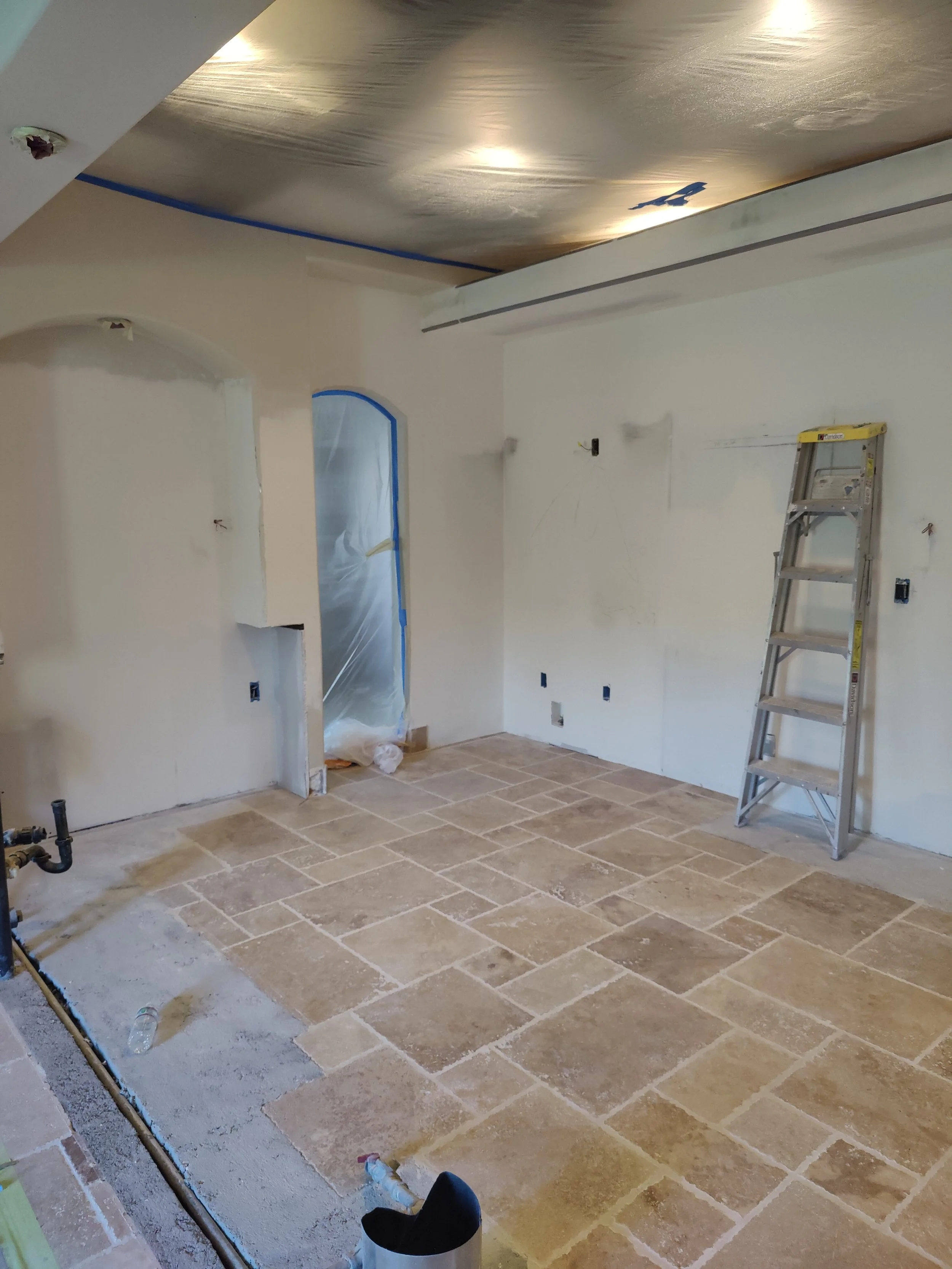 Room under renovation with beige stone tile flooring, a stepladder, and partially painted white walls. Plastic covering window and ceiling, construction materials scattered on the floor.