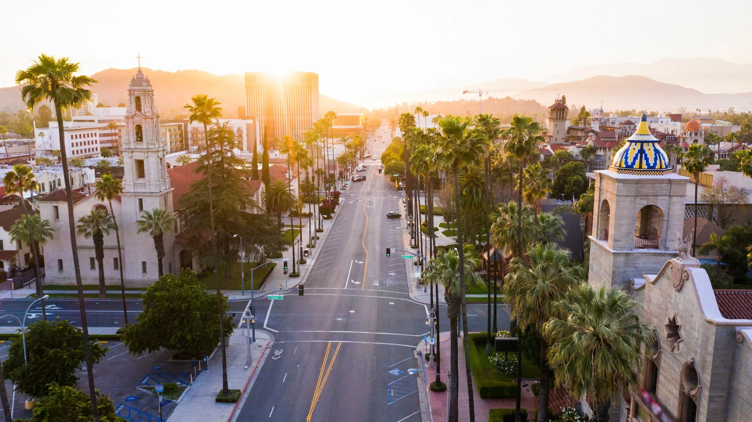 Downtown city street at sunrise with palm trees, historic buildings, and mountains in the background.