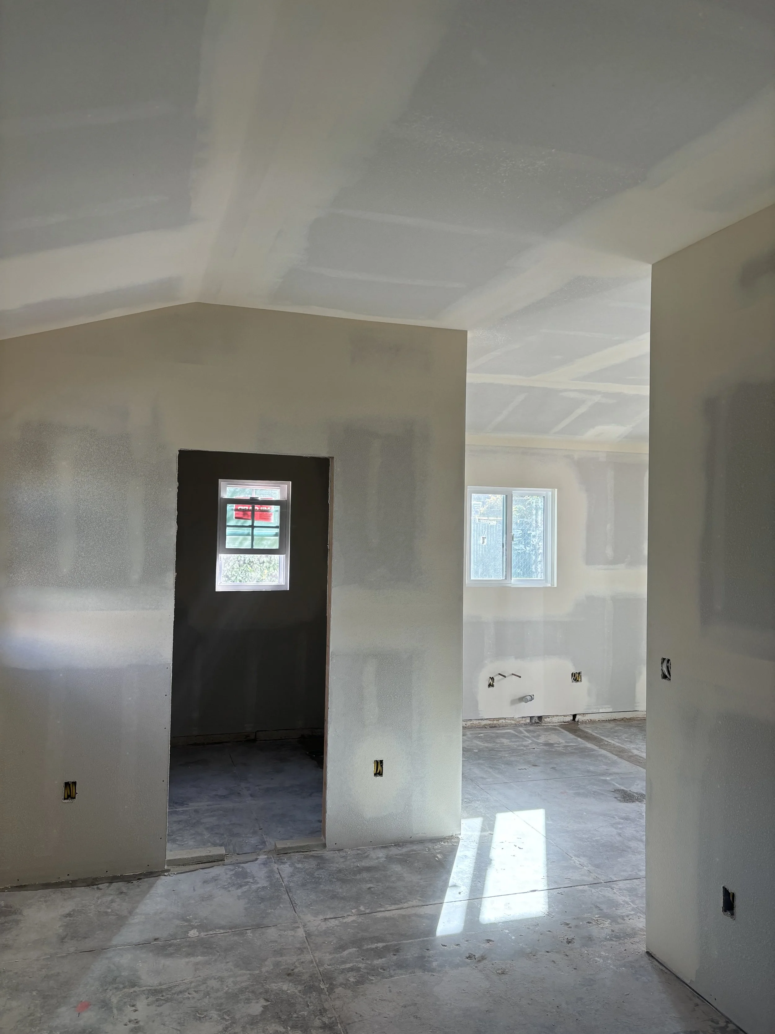 Interior of a house under construction with drywall partially installed, bare concrete floors, and small windows letting in natural light.