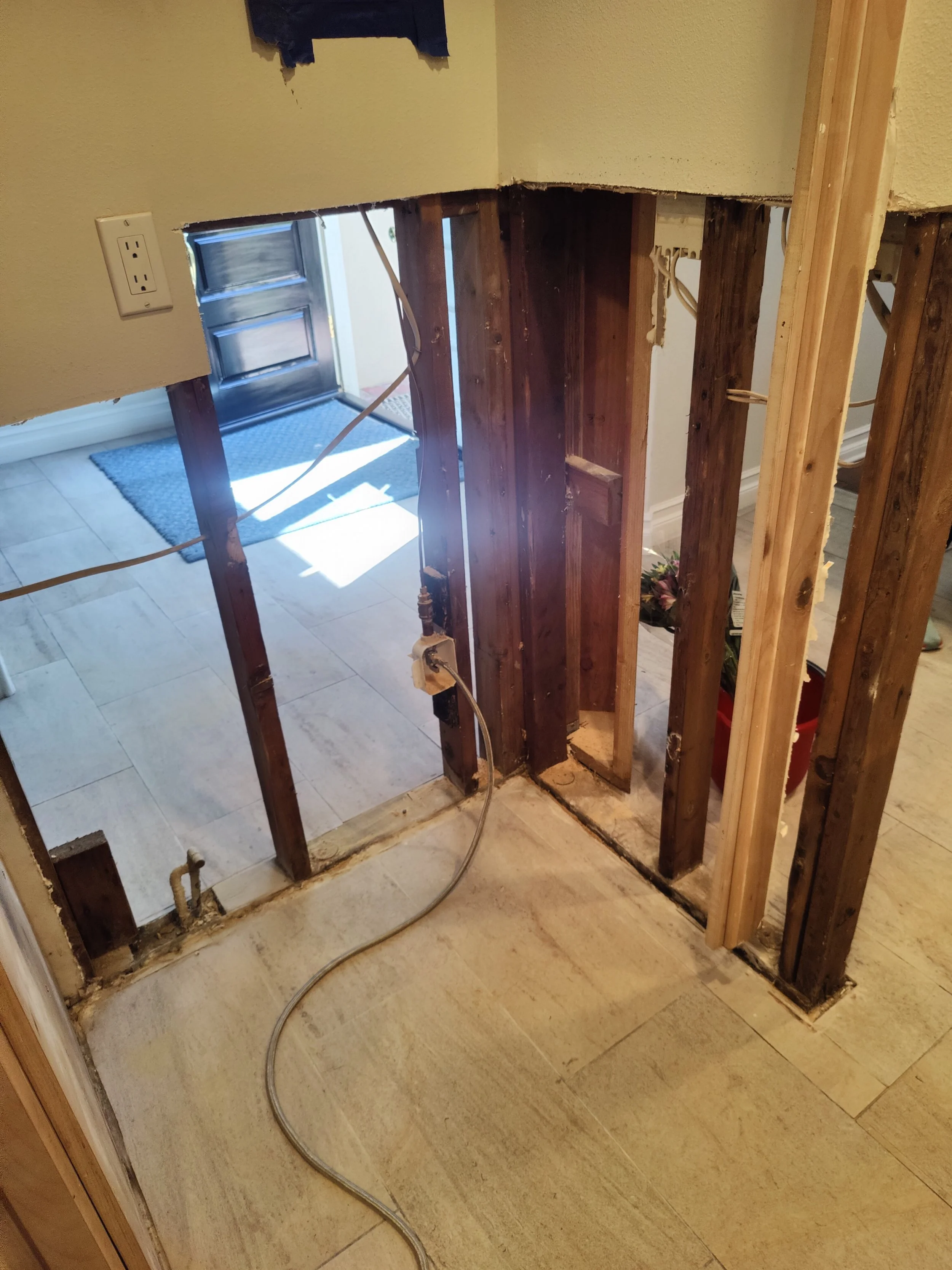 Bathroom wall with removed fixtures, showing exposed wooden studs and electrical wiring, with a view into the adjacent room with a door, mat, and tile flooring.