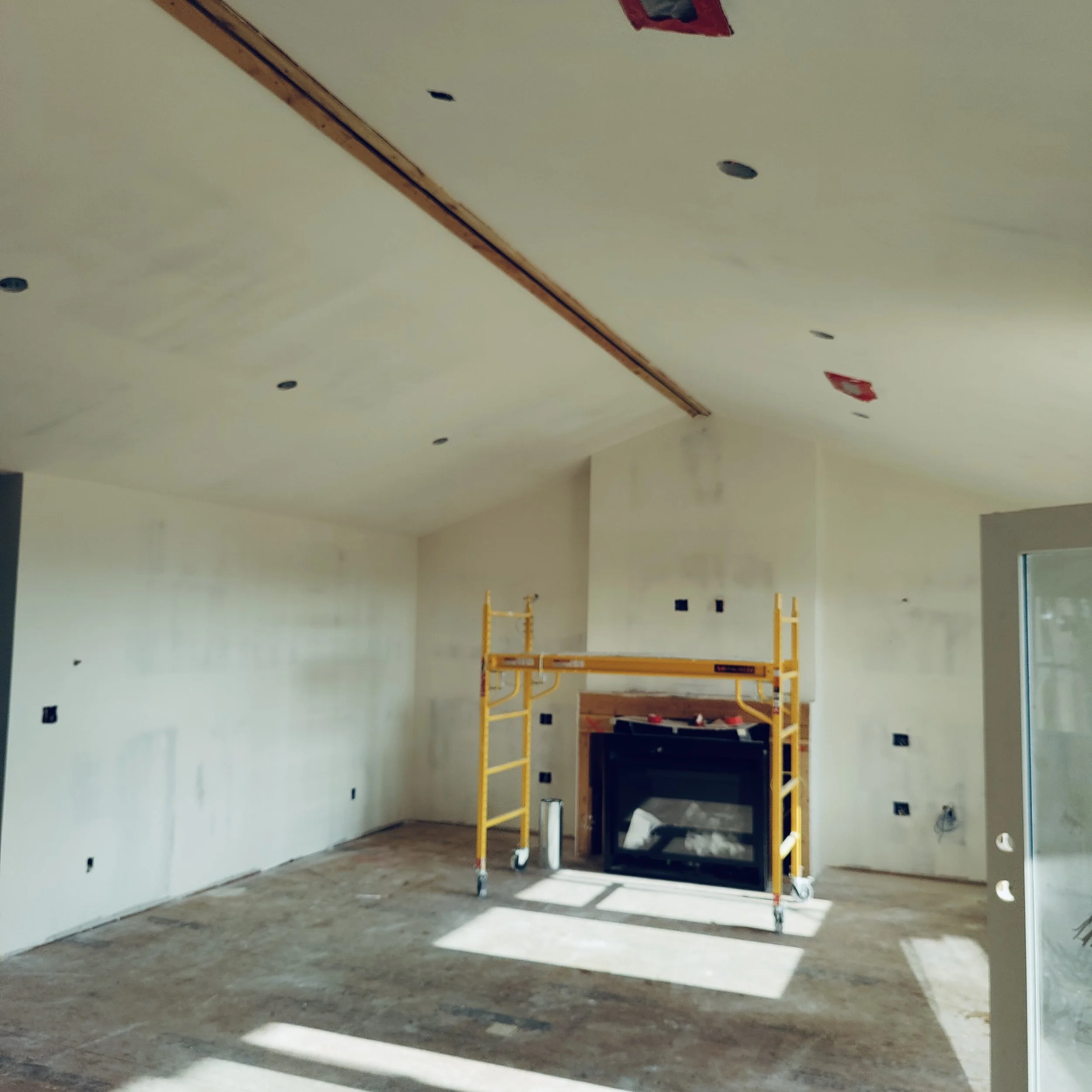Living room under construction with a yellow scaffold in front of a fireplace, unfinished walls, and a ceiling with cutouts for lights.