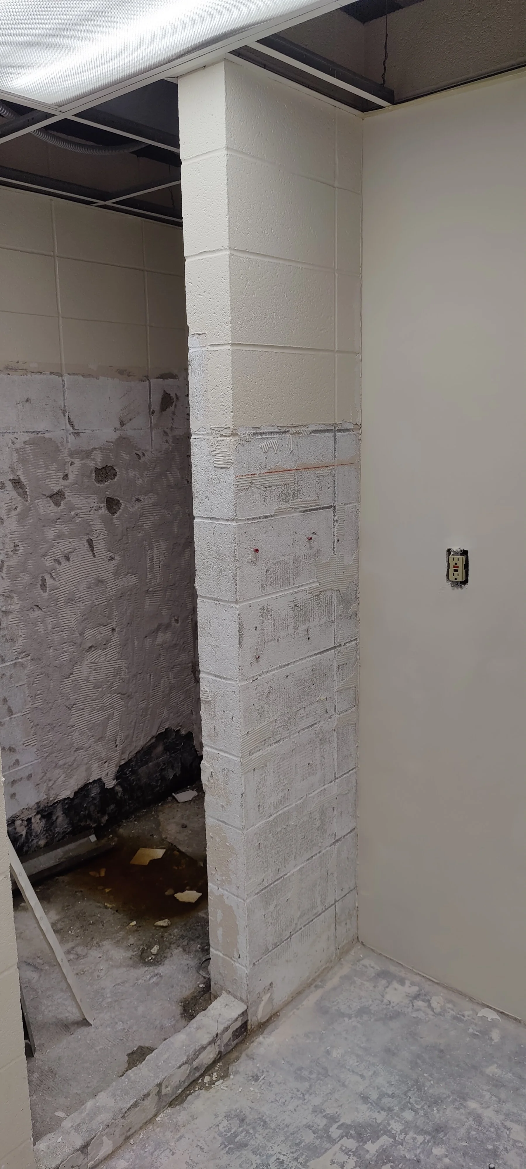 A partially constructed or renovated room with a white cinder block wall, a beige painted wall, and exposed ceiling with wiring. The floor has patches of concrete and construction debris.