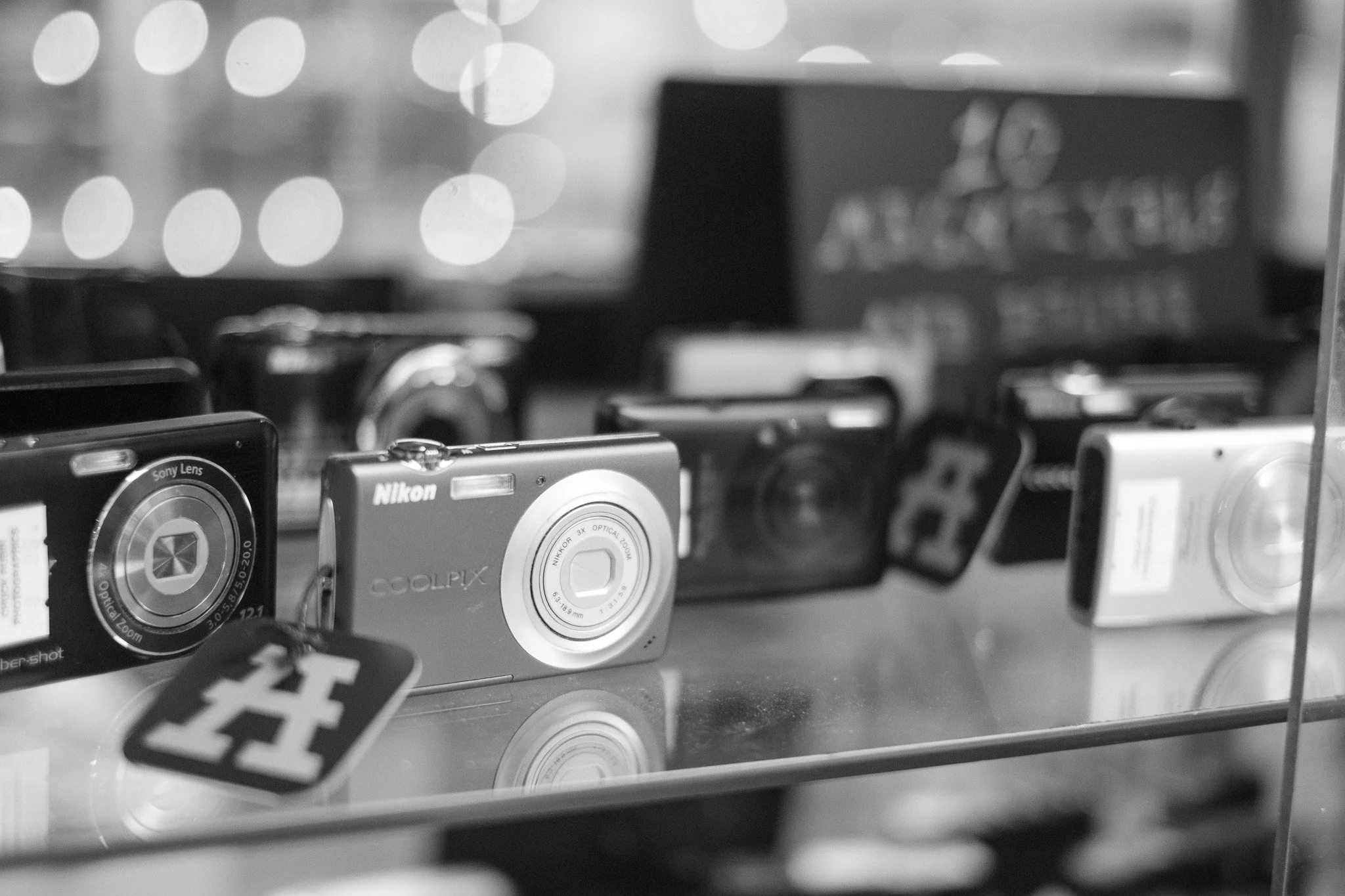 A display of various digital cameras on a glass shelf, including a Nikon Coolpix camera and a Sony Lens camera, with blurred lights in the background.