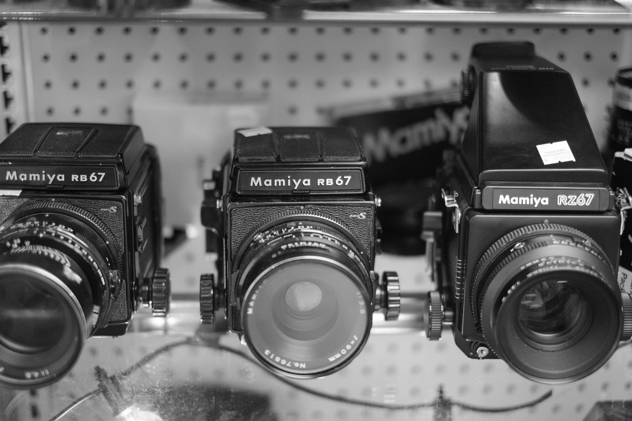 Three vintage Mamiya cameras displayed on a shelf in a store or collection, all with prominent lenses and model names visible.