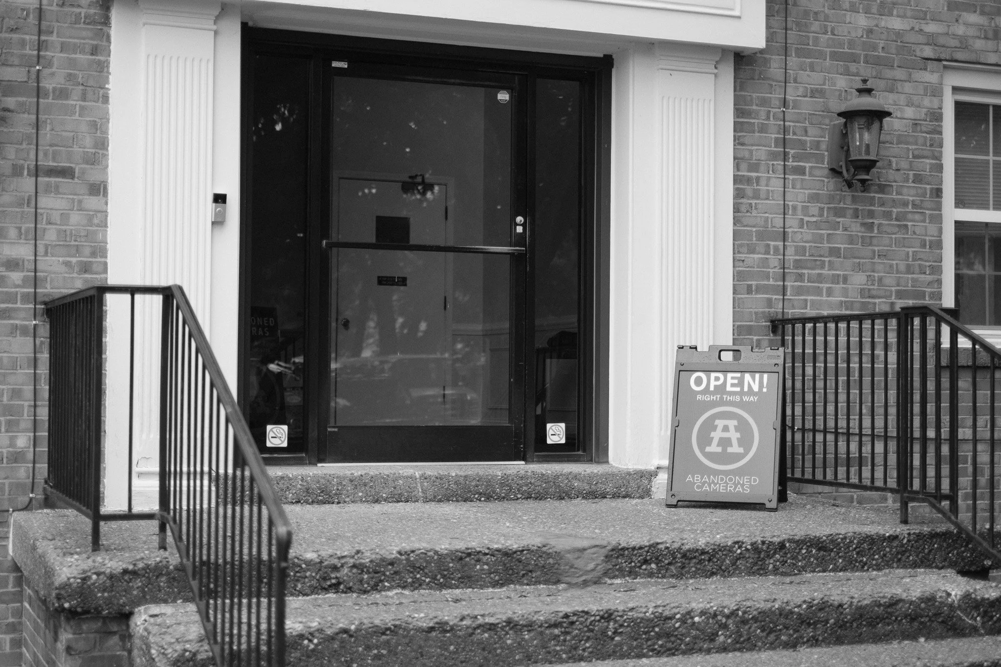 The entrance to a building with stone steps, black metal railings on both sides, a glass door, a sidewalk sign that reads 'Open! Right this way, Abandoned Cameras,' and a brick exterior wall with a wall-mounted lantern.