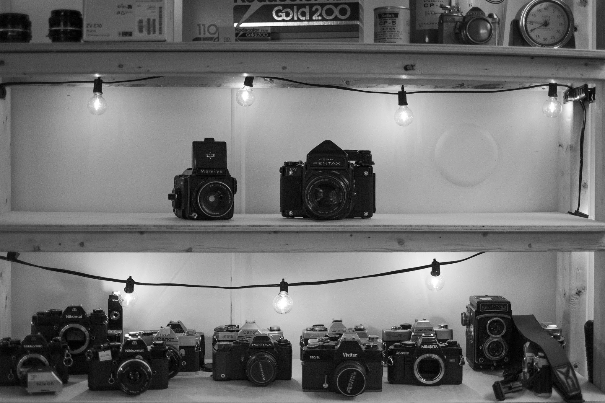 A wooden shelf displaying various vintage cameras, including two on the middle shelf and several more on the bottom shelf, with string lights hanging above.
