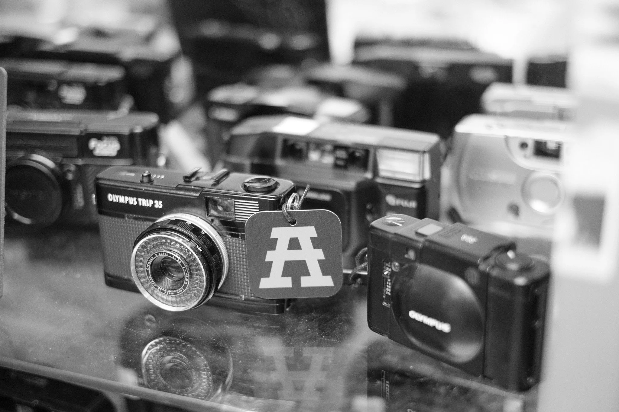 Several vintage Olympus film cameras displayed on a glass surface, with one camera having an 'A' logo tag hanging from it.