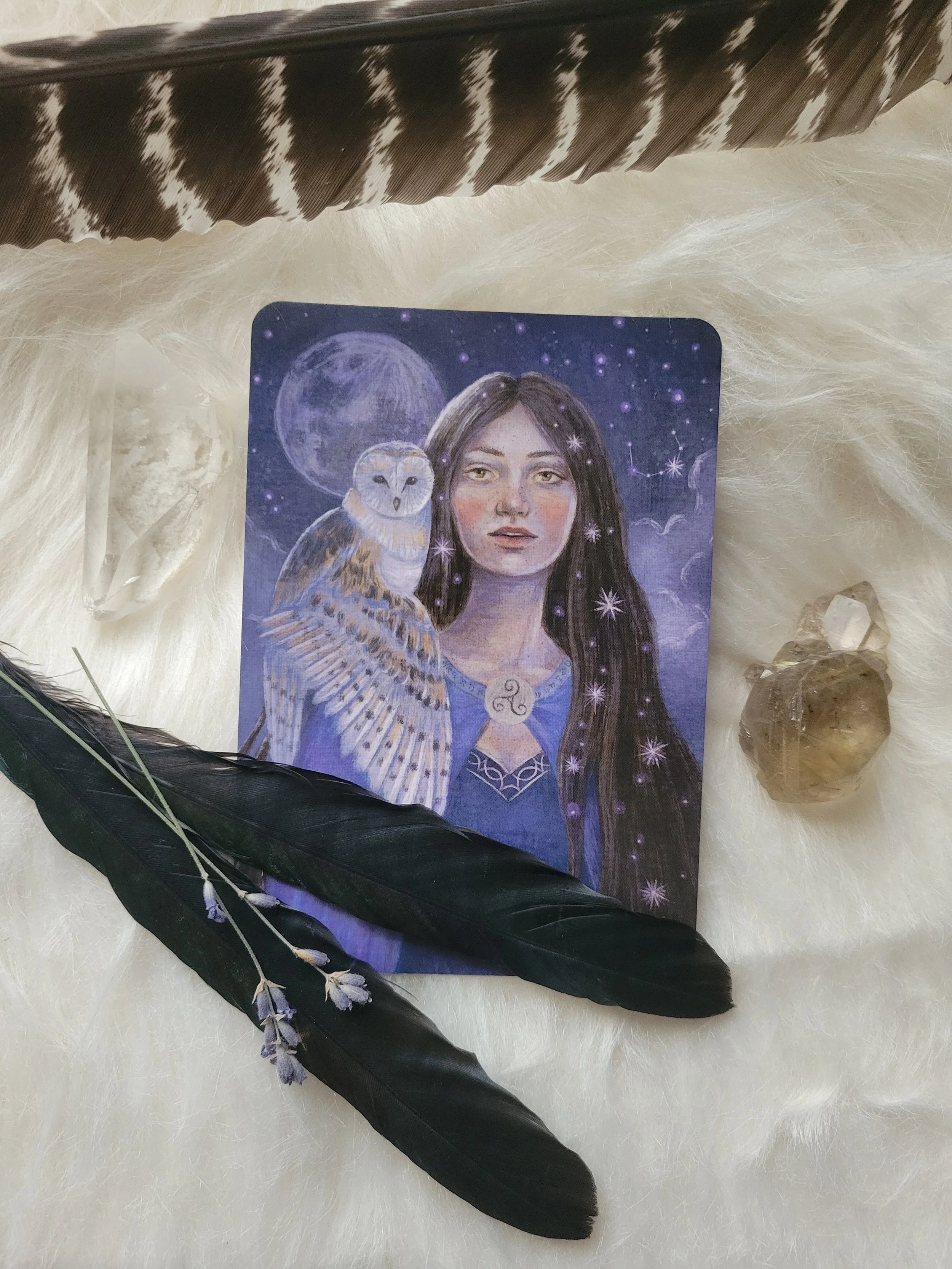 A mystical illustrated card featuring a woman with long dark hair, a moon behind her, an owl perched on her shoulder, surrounded by stars and constellations, with a large crystal and two feathers nearby.