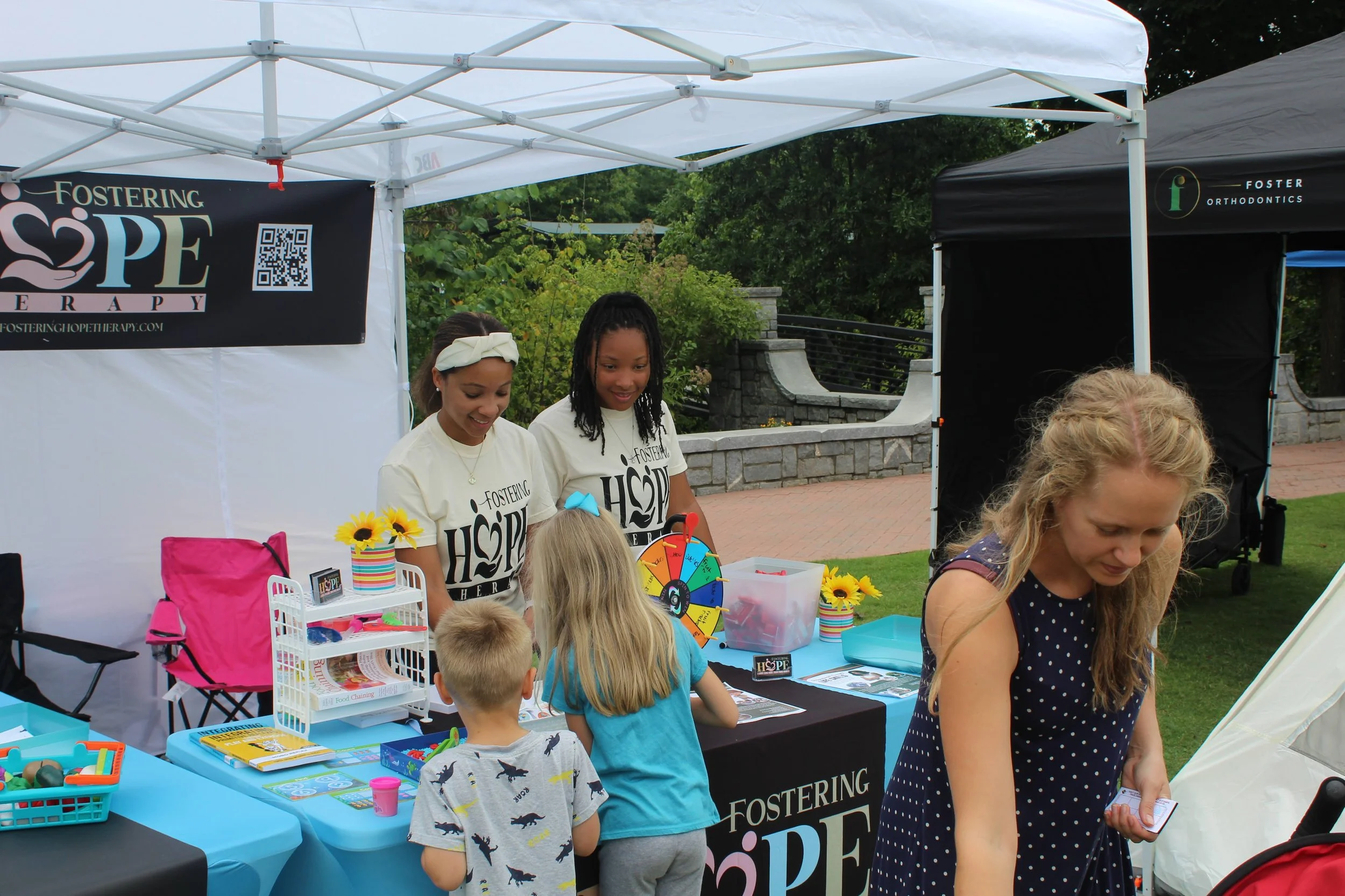 Children and two women at a community event booth under a white canopy, with a table holding informational materials, a colorful spinning wheel, and sunflowers in a striped vase.