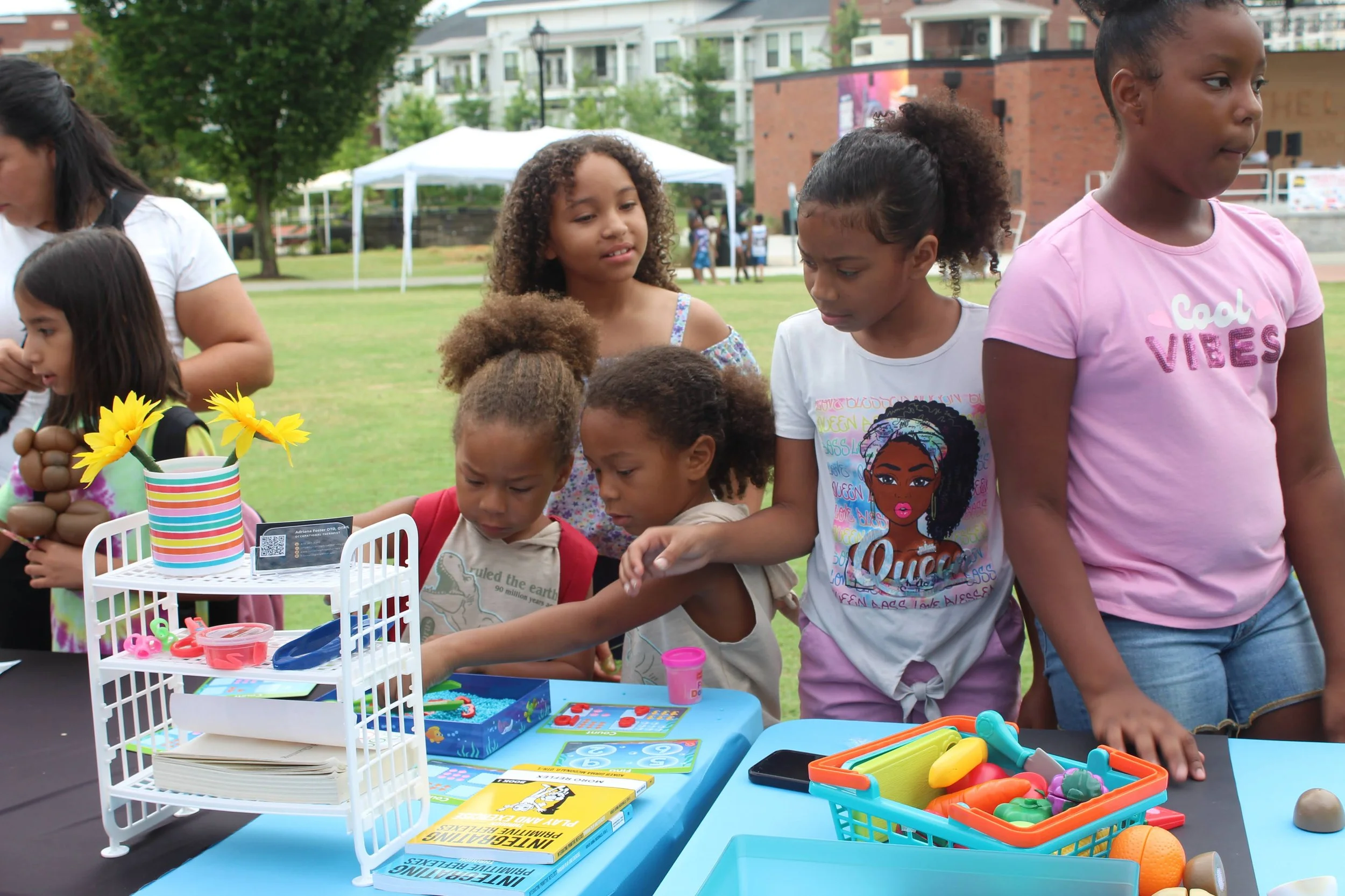 Group of children looking at toys and books on a table at an outdoor event in a park.