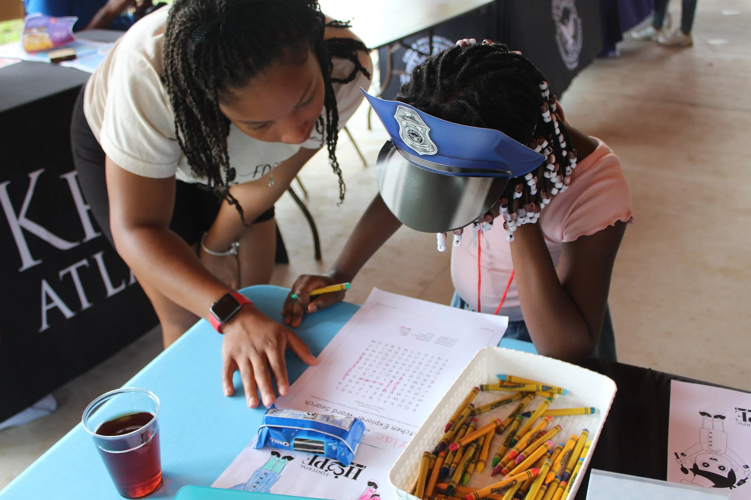 A woman and a young girl are working together at a table. The woman is leaning over, pointing at a worksheet, while the girl covers her face with her hand, wearing a police cap. The table has crayons, a glass of tea, and some papers on it. In the bac