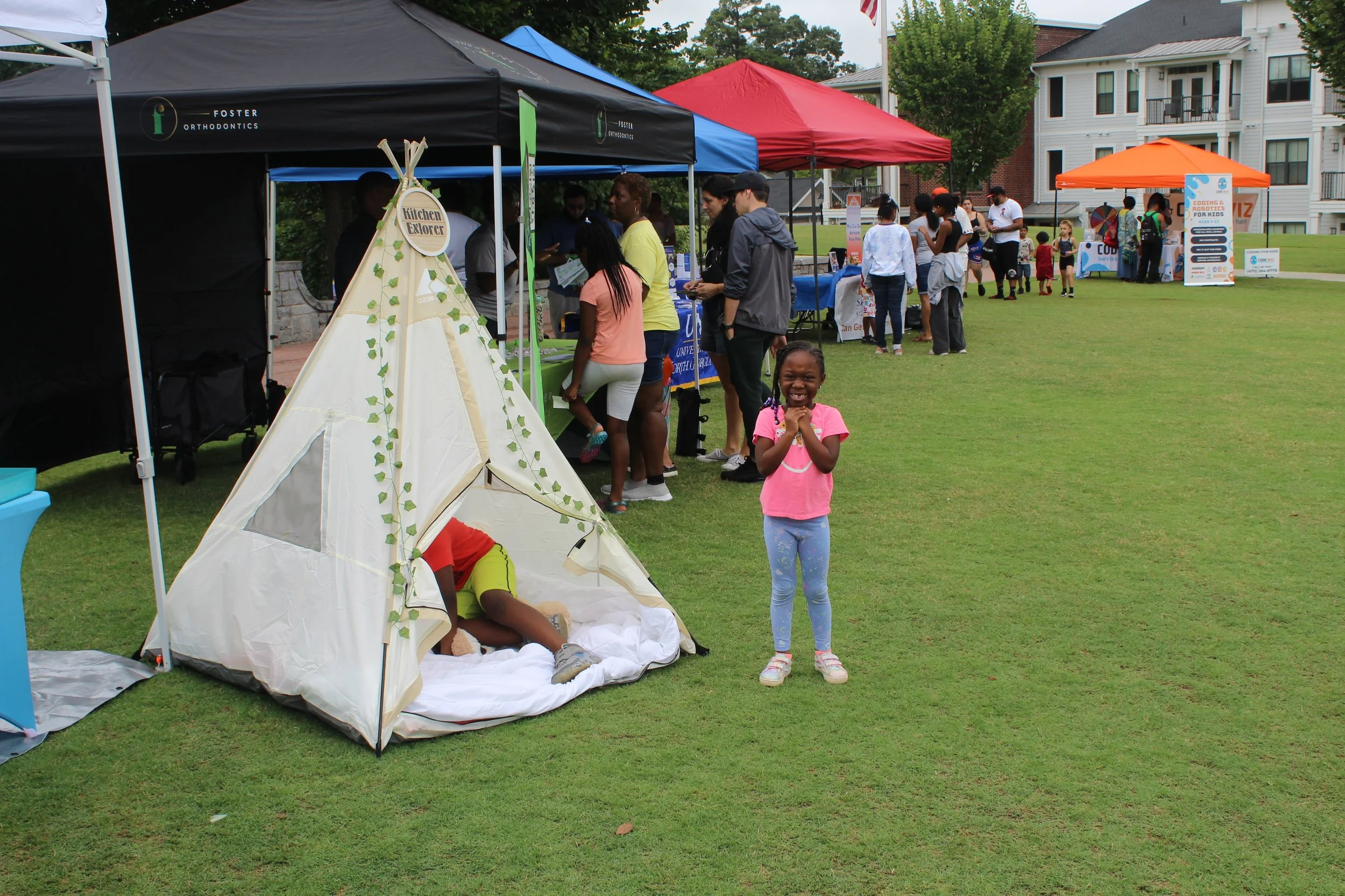 A young girl in a pink shirt and blue leggings smiling with hands near her face, standing next to a small pretend teepee tent on a grassy field during an outdoor community event. People are gathered around tents and booths in the background.