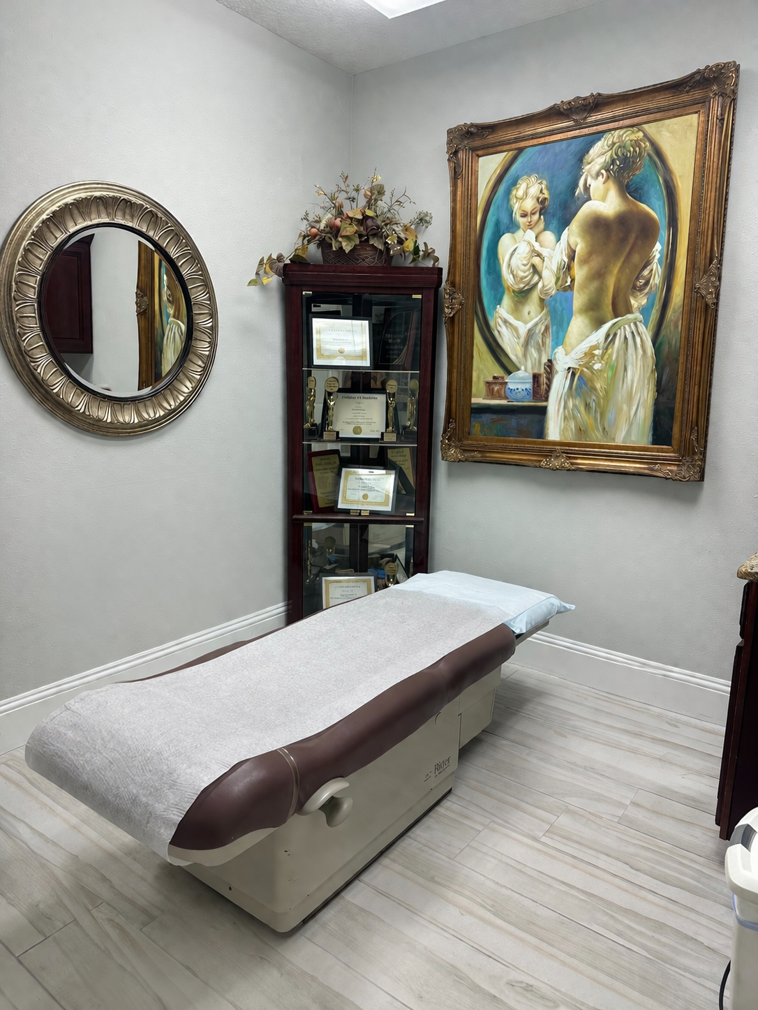 A treatment table in a procedure room for BHRT (bio-identical hormone therapy) with a large painting on the wall, a decorative round mirror, and a display cabinet with awards and certificates.