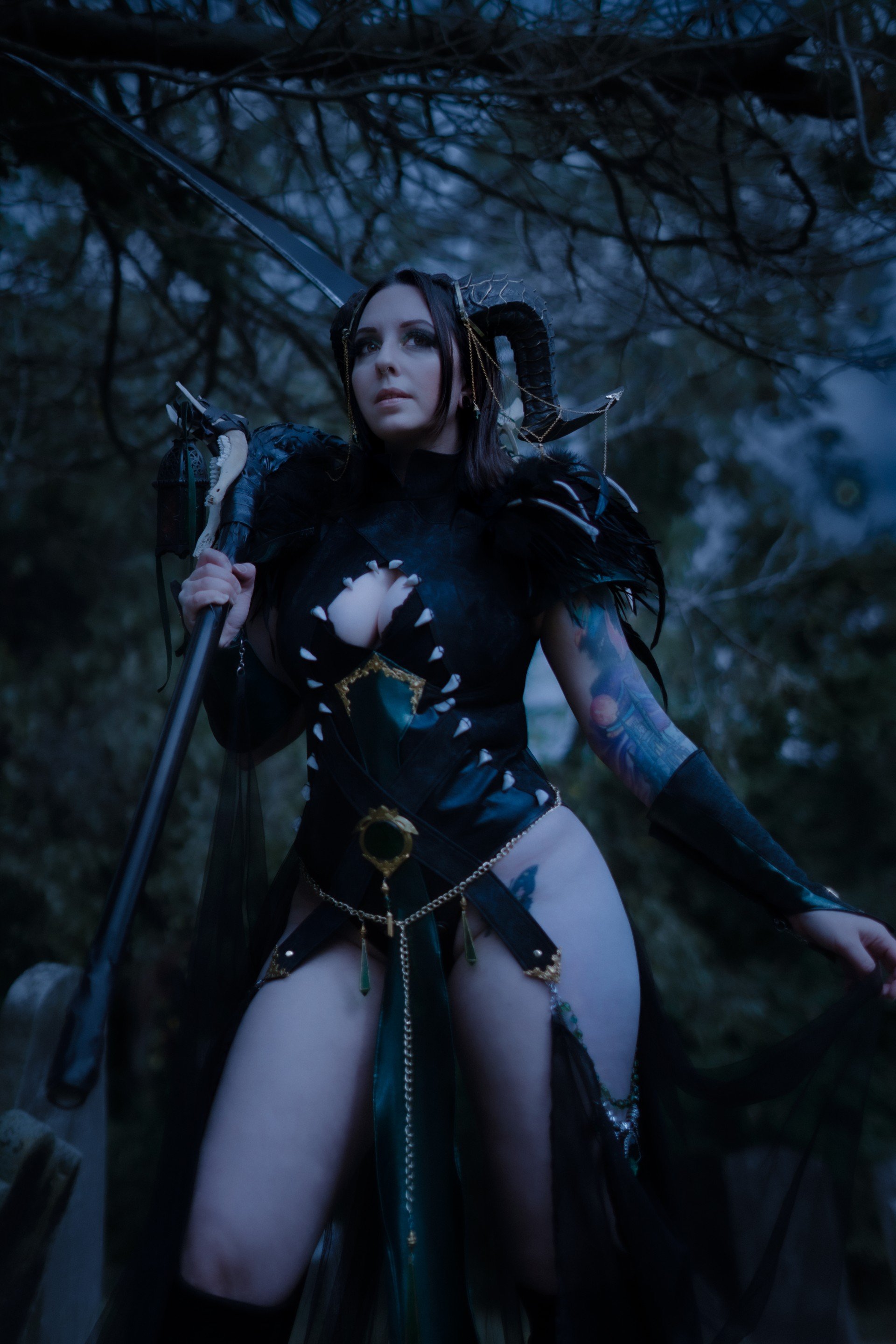 A woman dressed as a fantasy warrior with dark hair, horned headpiece, and black armor, holding a sword in a dark forest at dusk.