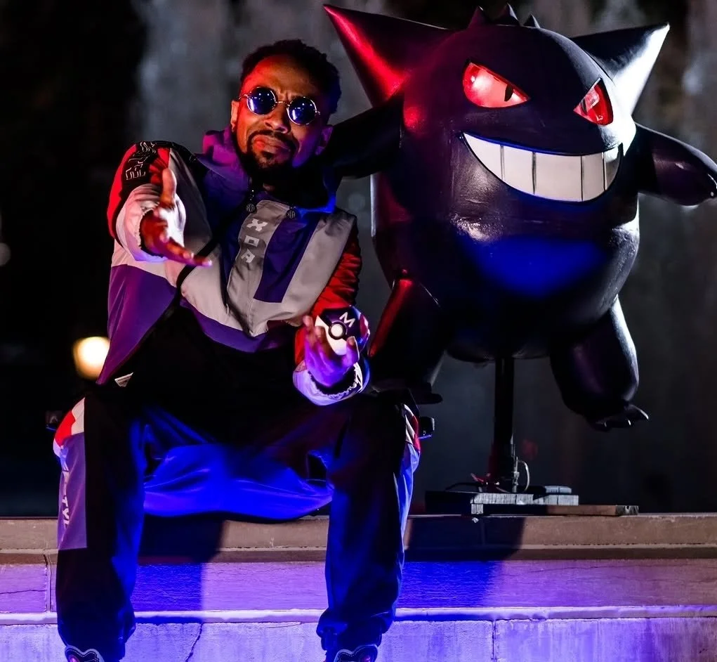 A man wearing sunglasses and a colorful jacket in a crouched pose next to a large inflatable Pokémon, Gengar, with glowing eyes and a big smiling mouth.