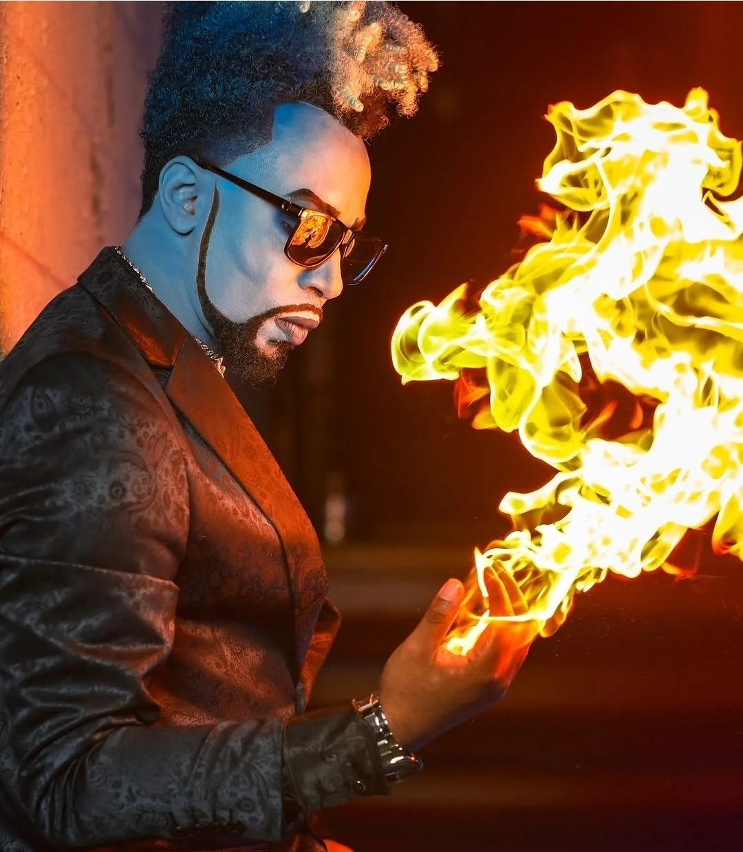 Man wearing sunglasses and a dark patterned jacket looking at flames coming from his hand in a dark setting.