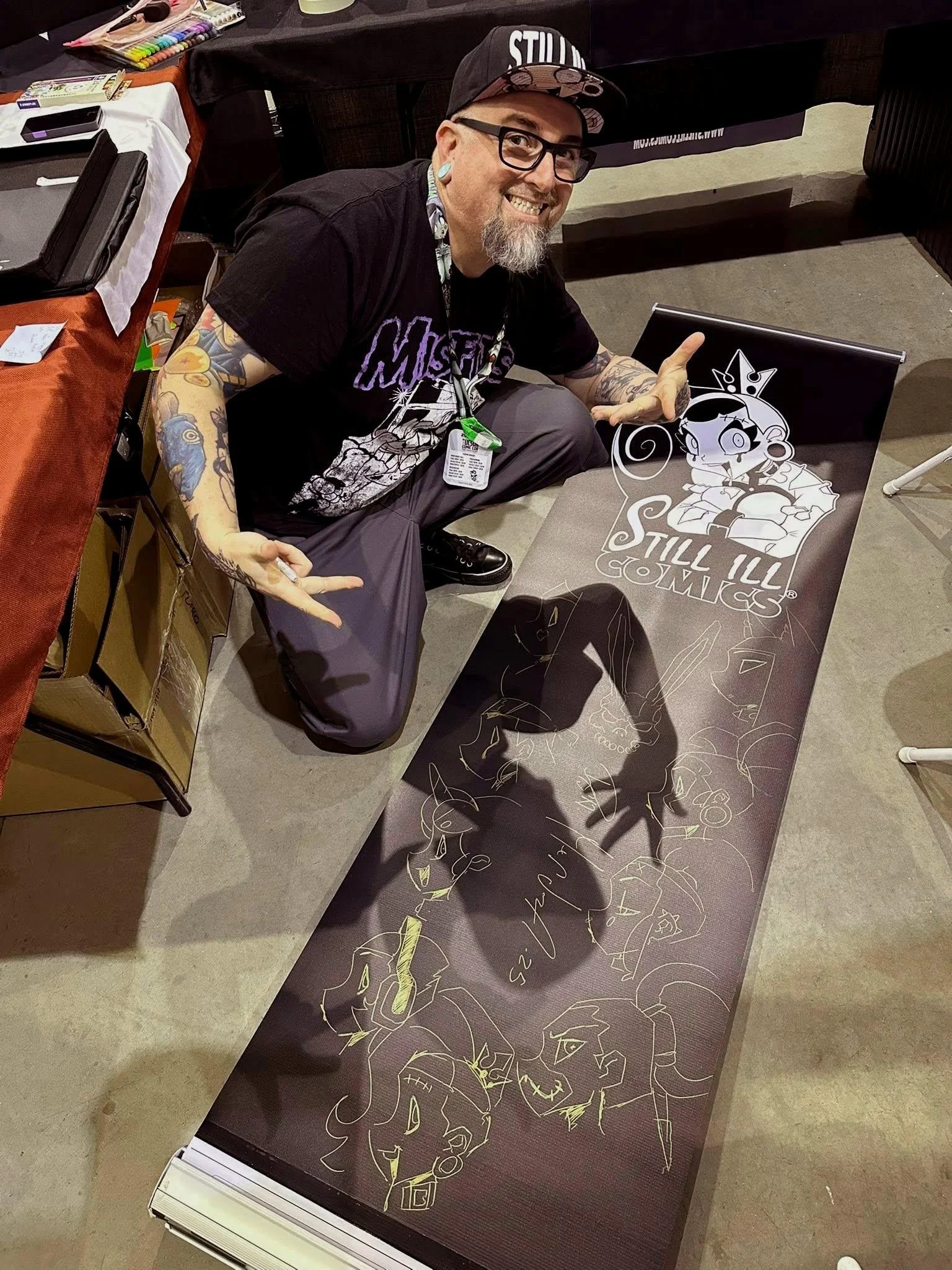 A man with glasses, tattoos, and a beard is crouched next to a large signed art print of cartoon characters, at a convention table. He is smiling and pointing at the artwork.