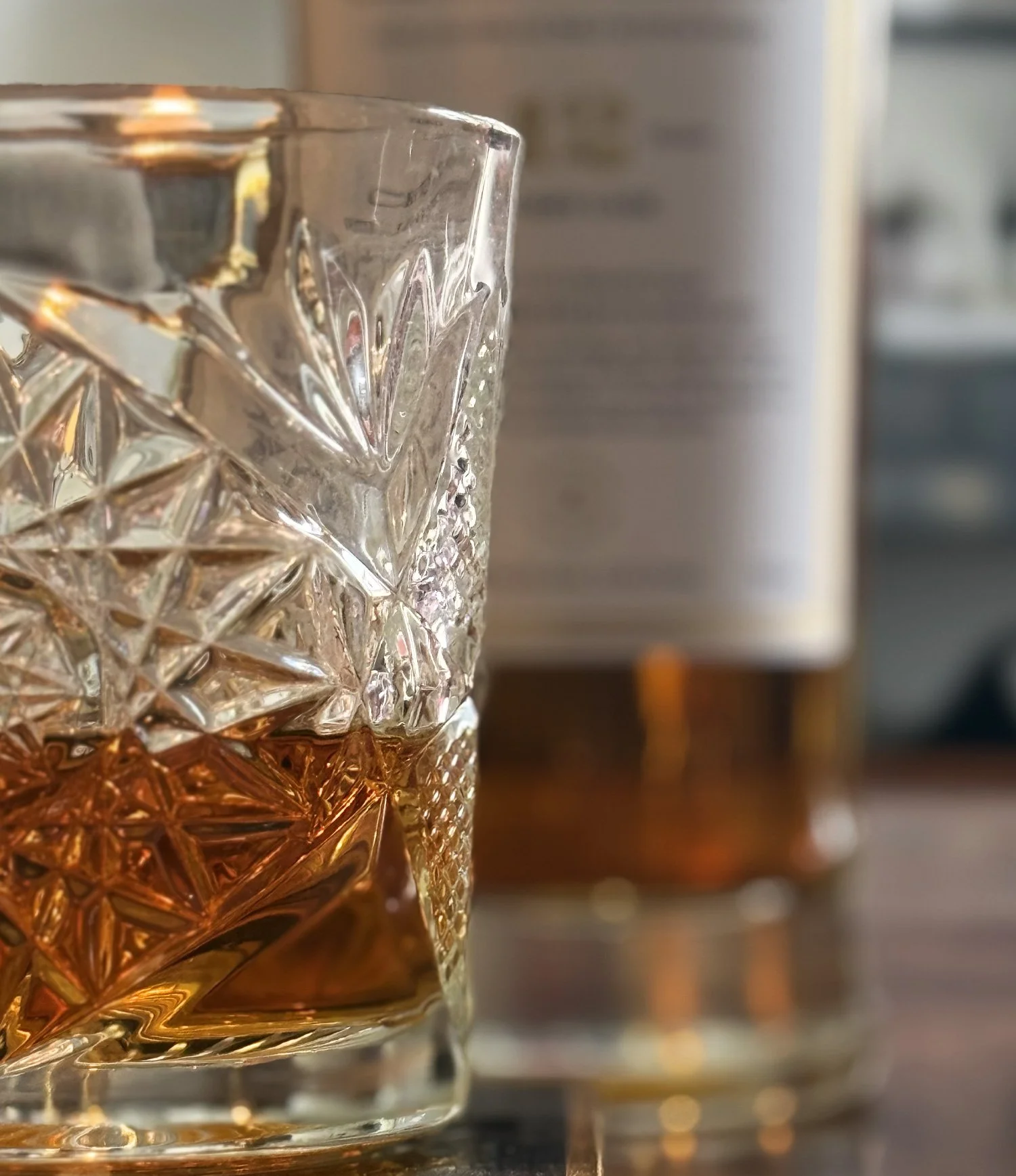 Close-up of a decorative glass with a detailed, cut pattern, filled with a golden-colored liquid, placed on a dark surface with a blurred background of Glen Loch 12 Years Single Malt Scotch.