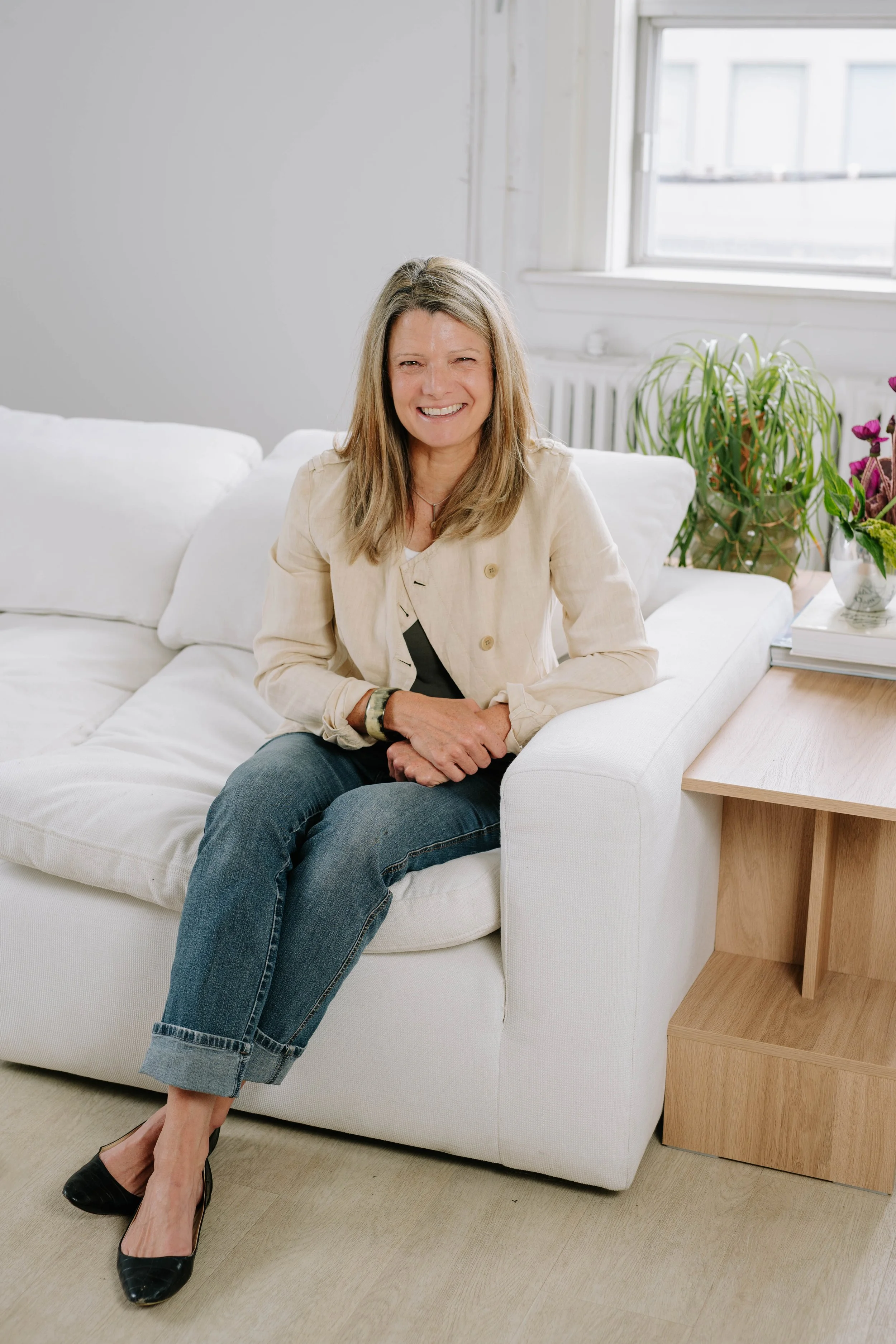 A woman sitting on a white couch in a bright room, smiling at the camera. She has shoulder-length blonde hair and is wearing a beige blazer, black shoes, and jeans. There are green plants and flowers on a side table next to her, and a window with natural light coming through in the background.