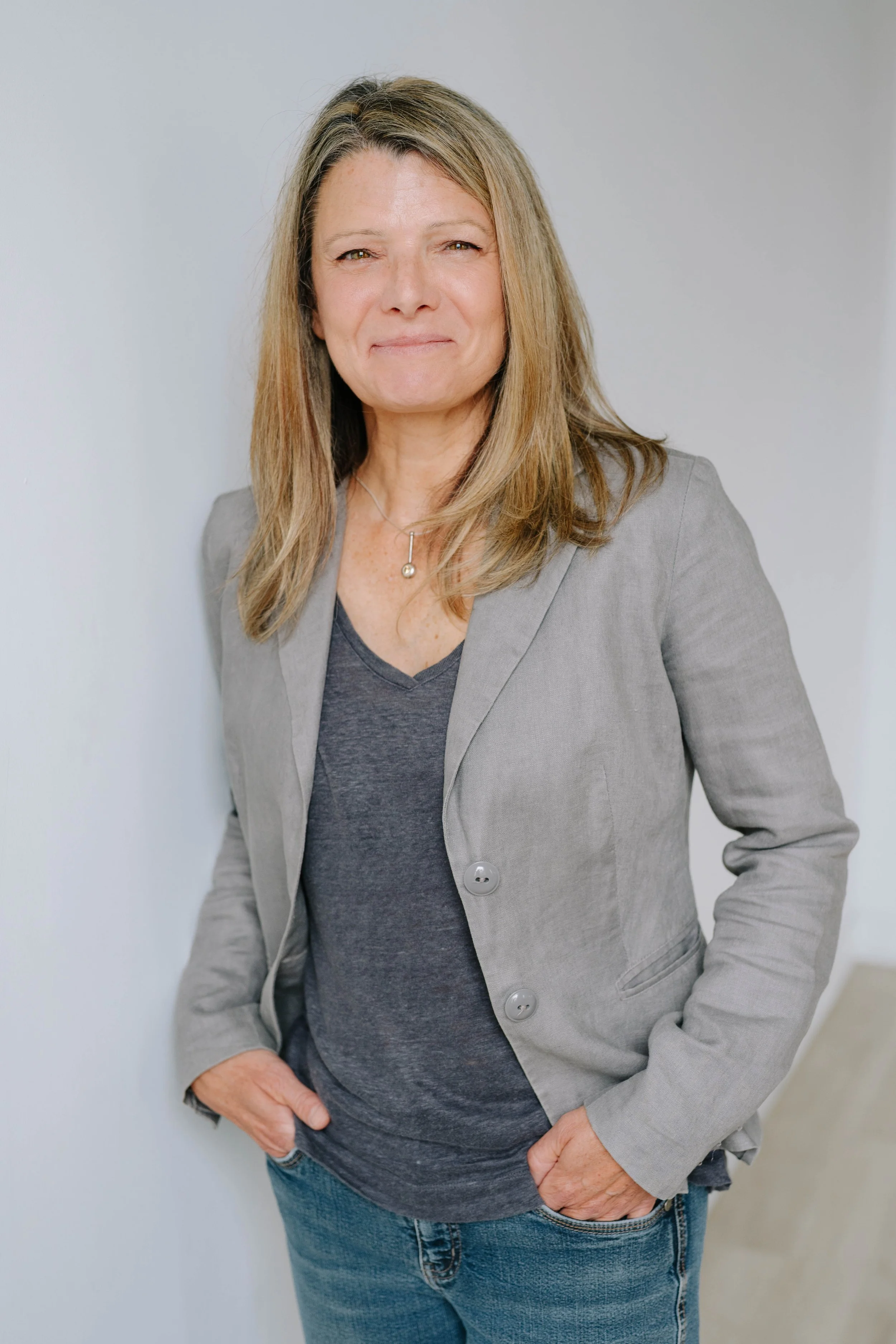 Woman with shoulder-length blonde hair wearing a grey blazer over a dark grey t-shirt, standing against a plain light-colored background.